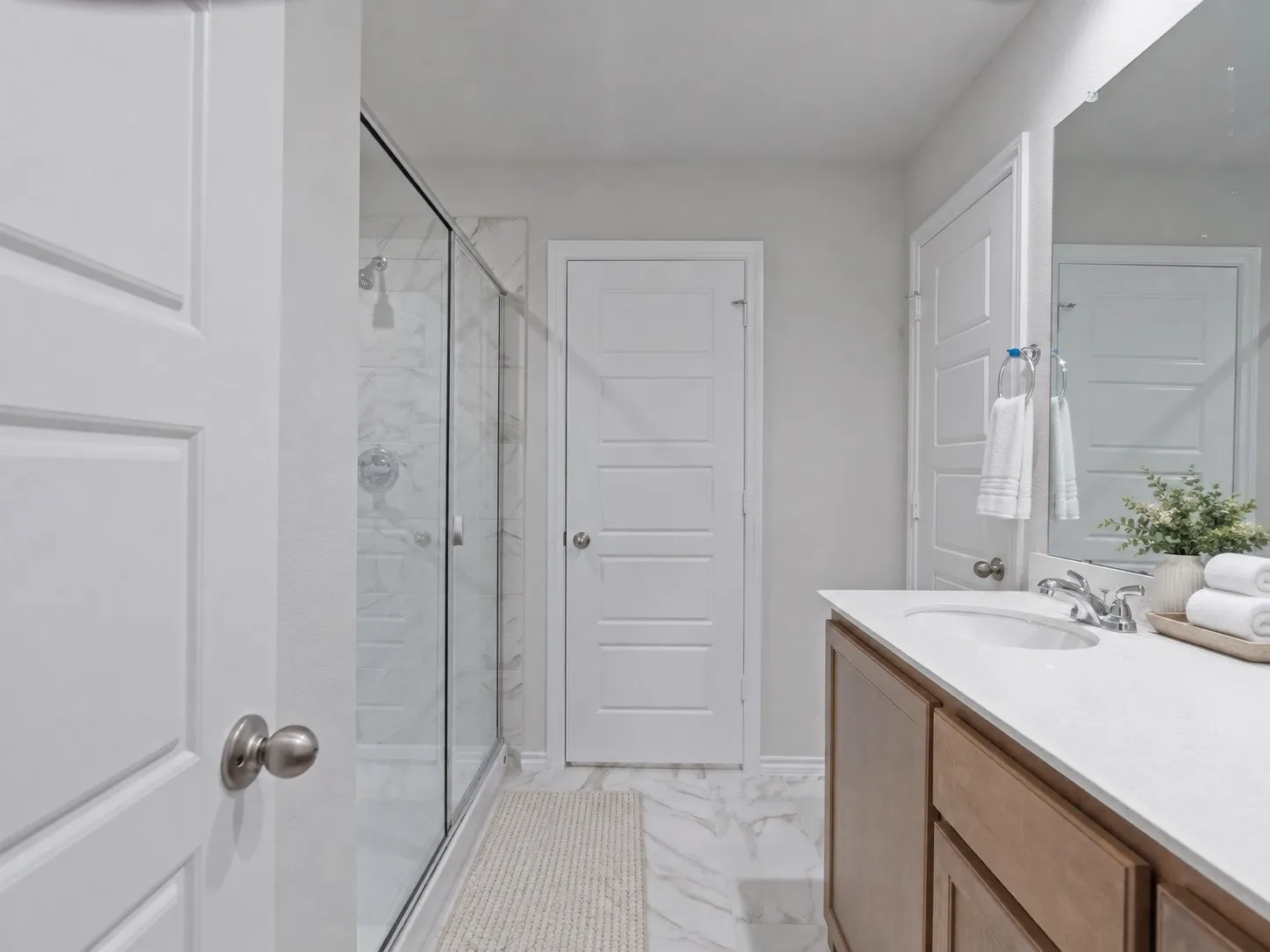 Walk-in shower with clear glass enclosure, white stone-look tile flooring, wood-finish vanity with an integrated sink, and a large wall-mounted mirror