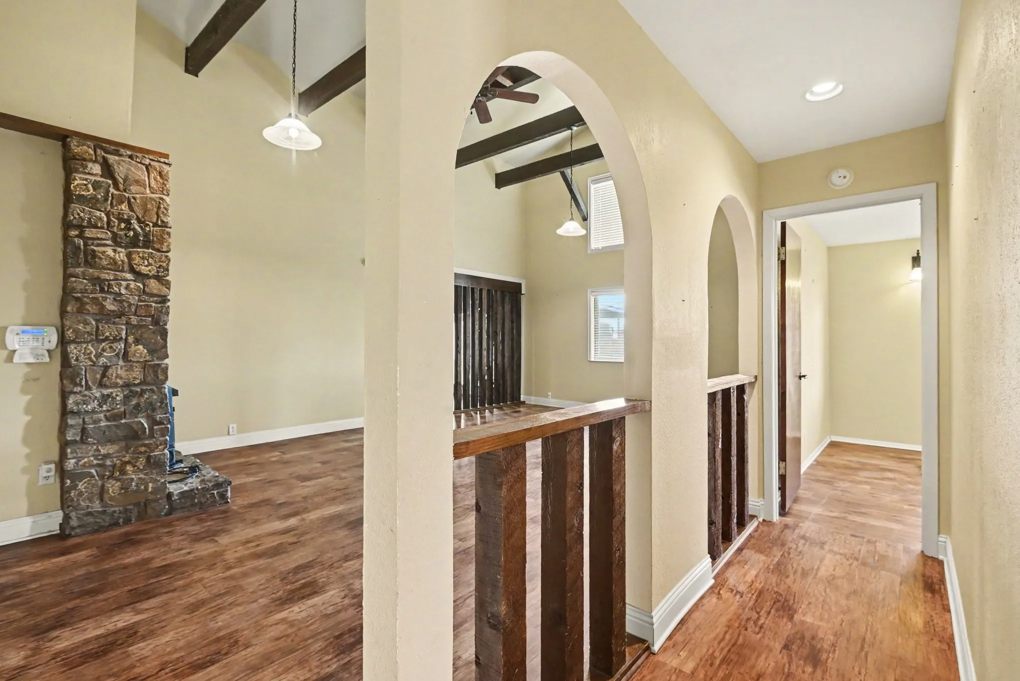 Vaulted ceiling with exposed beams, stone-clad fireplace, wood-finish flooring, interior archways, and a wooden banister