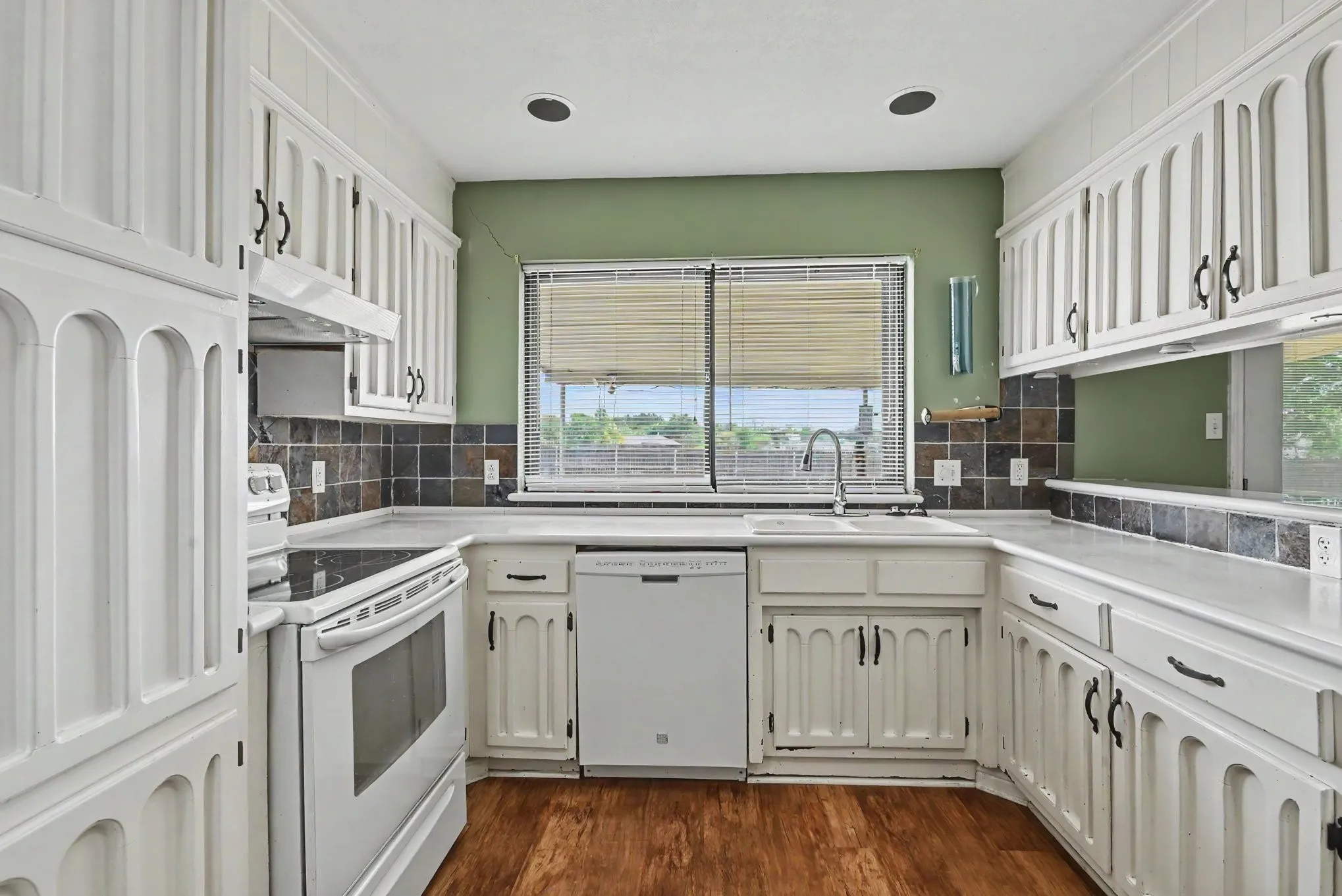 White cabinetry with decorative arch panels, dark tile backsplash, wood-finish flooring, and white appliances