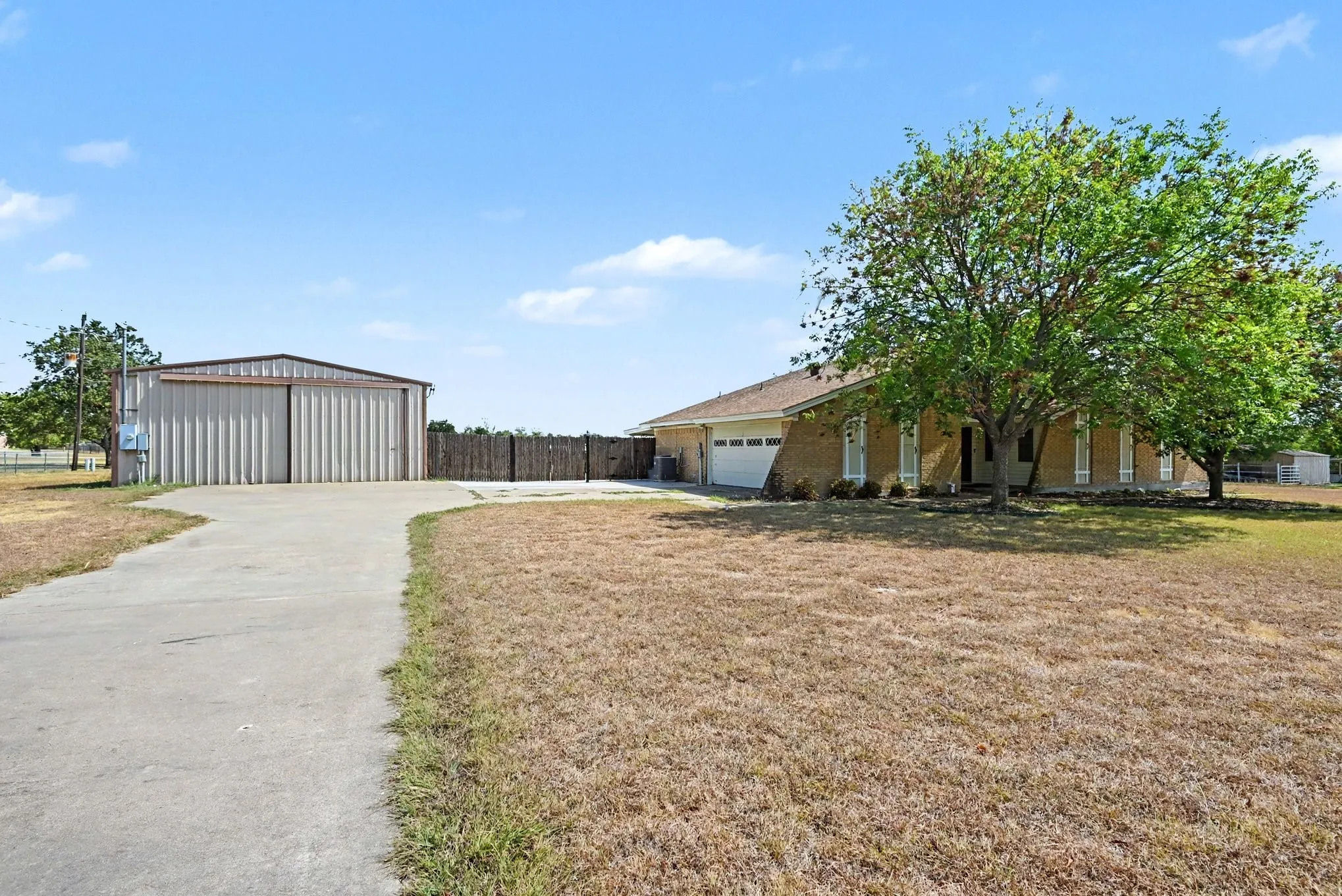 Brick exterior home with an attached garage, a separate metal building, a concrete driveway, and established trees