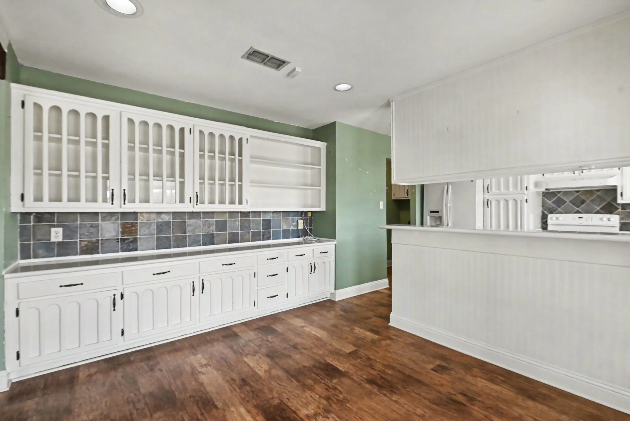 Expansive white built-in cabinetry with glass-front uppers and dark hardware, tile backsplash, wood-finish flooring, and a beadboard-paneled half wall