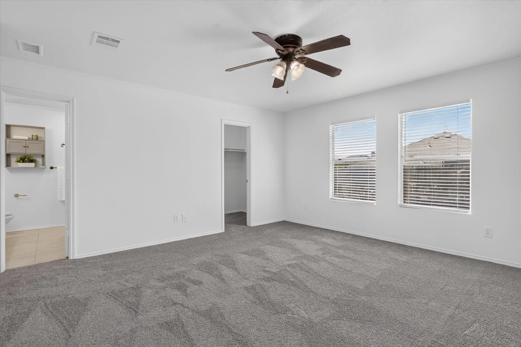Spacious room with gray carpeting, white walls, two windows with blinds, and a ceiling fan with light fixture