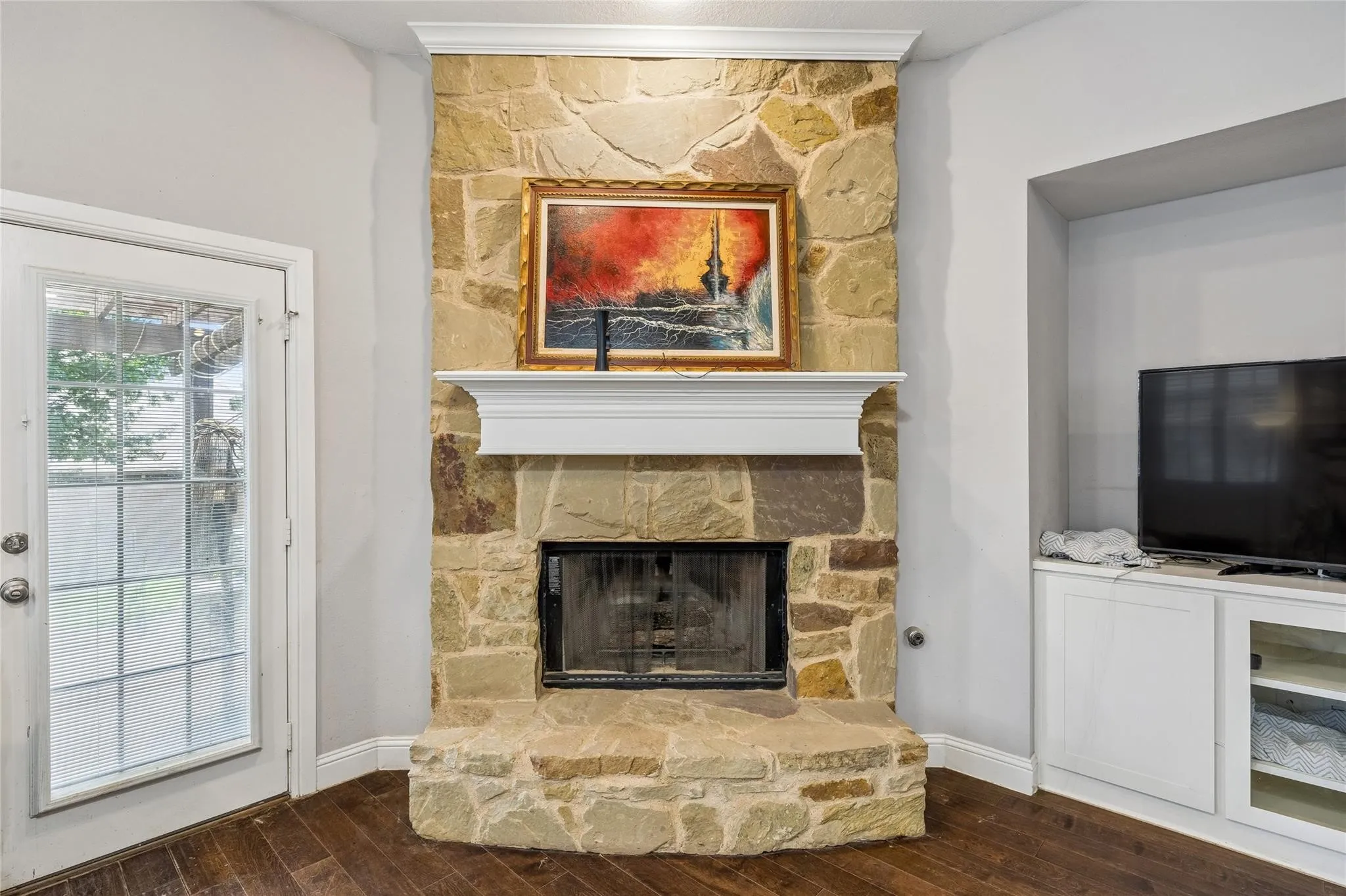 Stone-clad fireplace with a white mantel, built-in media cabinet with open shelving, dark wood-finish flooring, and a glass-paned door