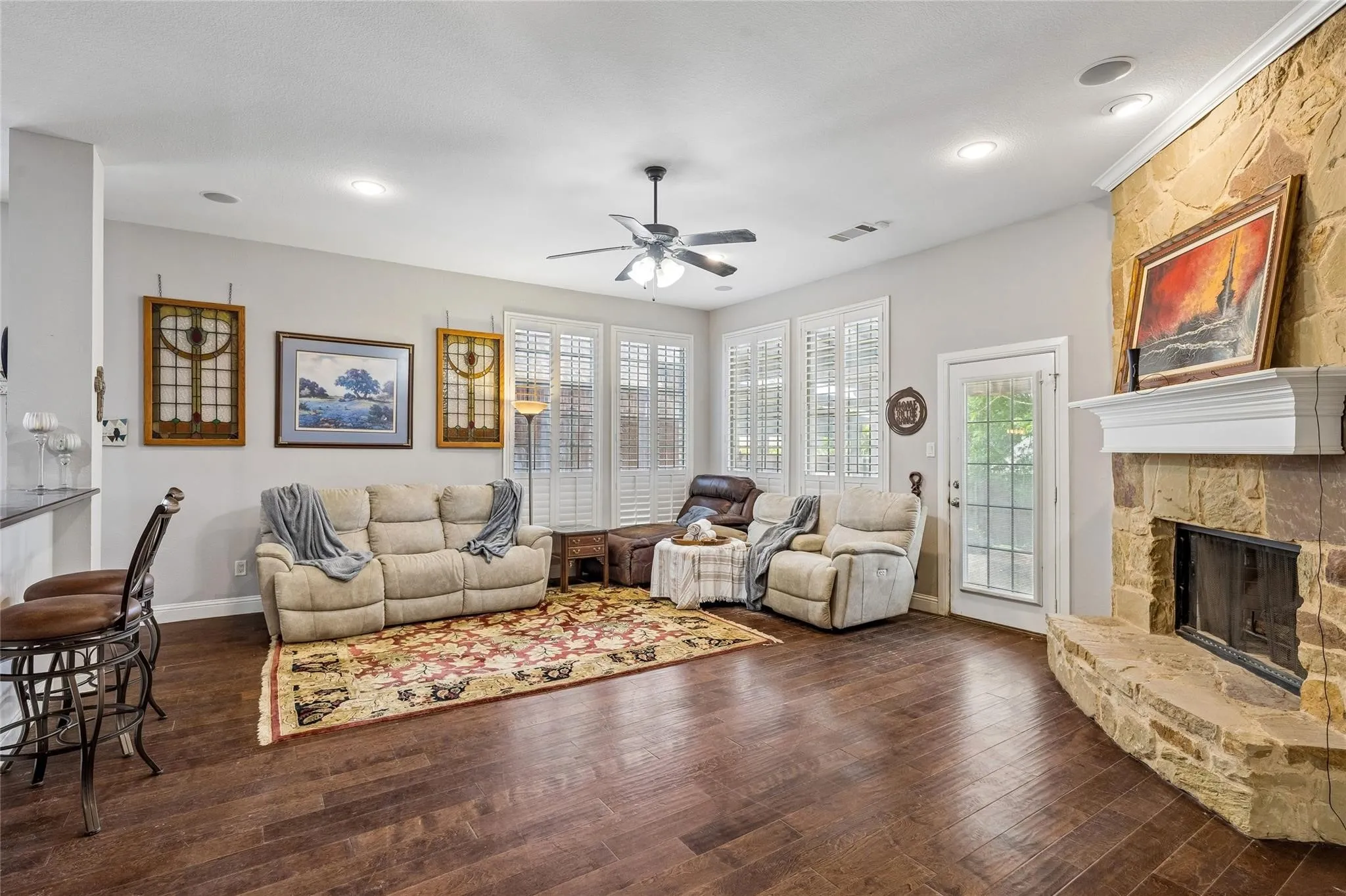 Stone-clad fireplace with mantel, hardwood flooring, ceiling fan, recessed lighting, and multiple windows with plantation shutters