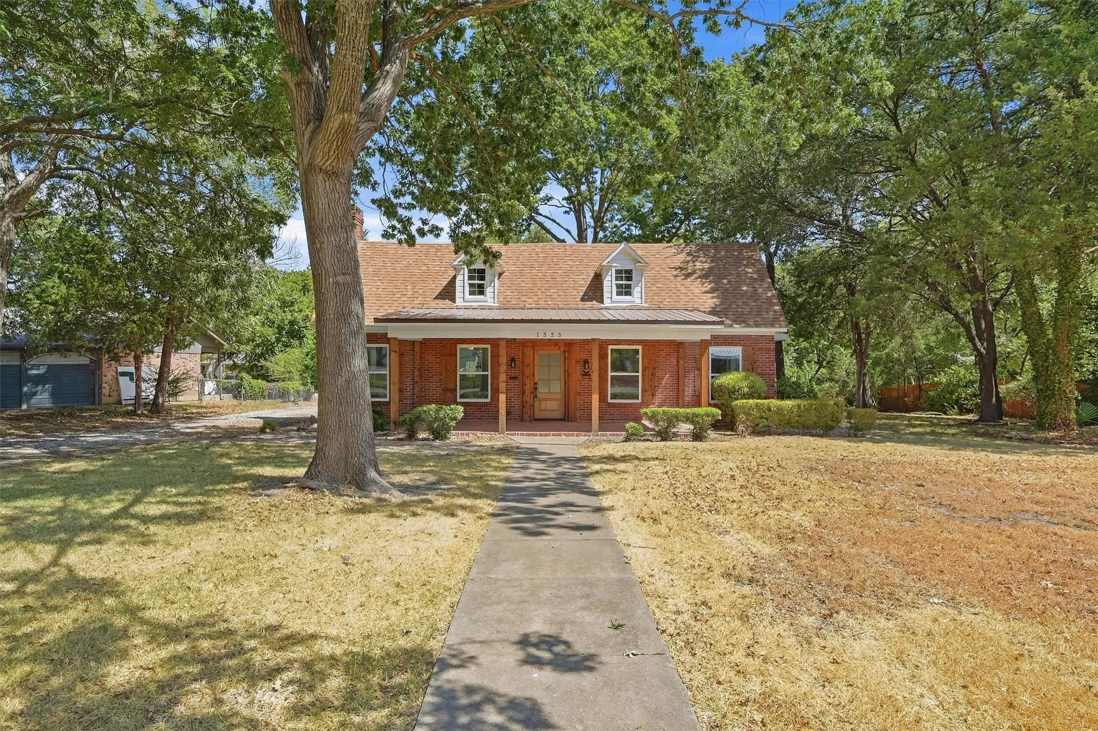 Brick exterior residence with a dormer roof, concrete pathway, and mature trees