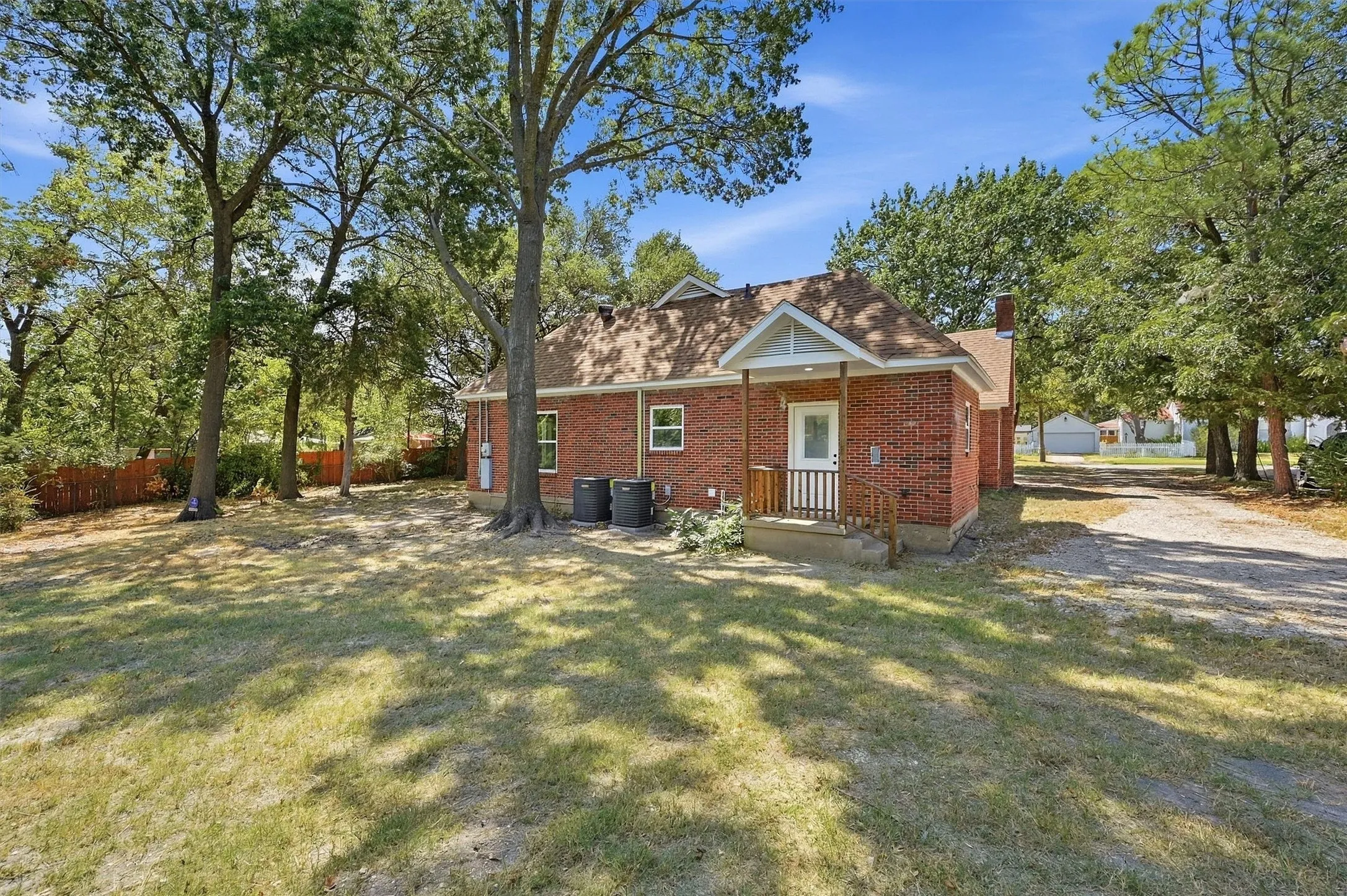 Red brick exterior, covered porch with white columns, mature trees, gravel driveway, and a shingle roof