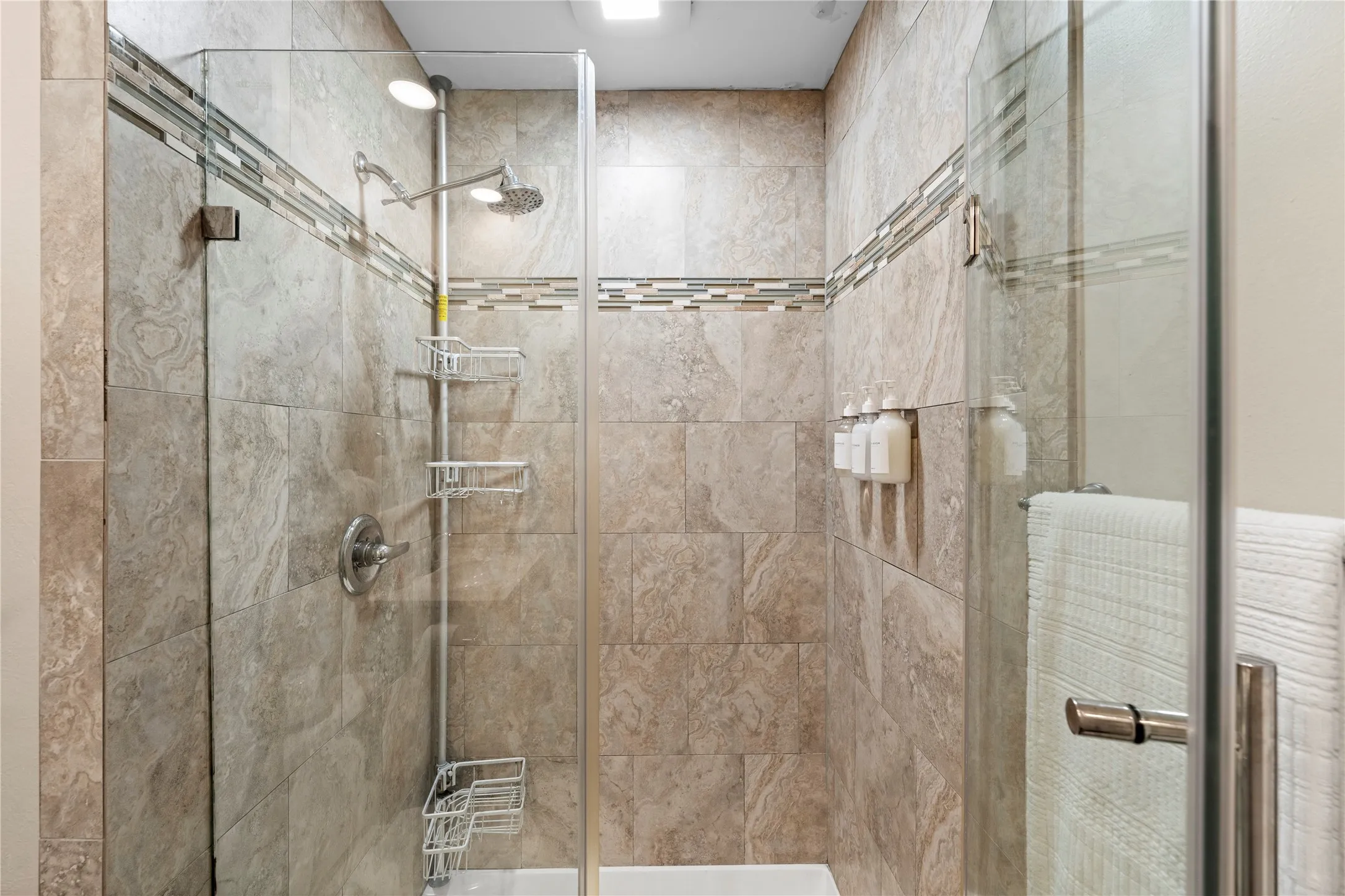 Glass-enclosed shower with a rainfall showerhead, handheld sprayer, and tiled walls featuring a decorative mosaic border
