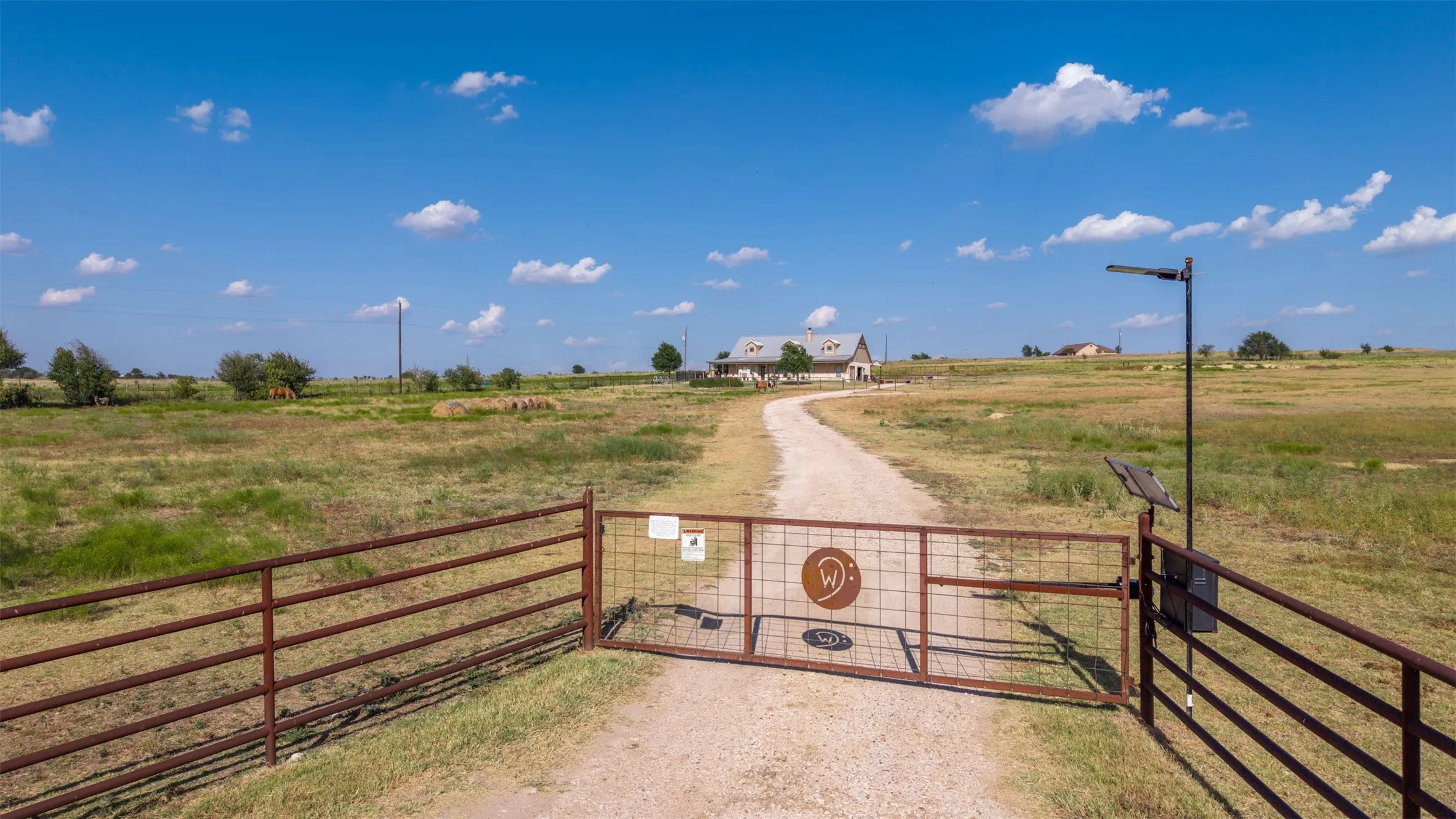 Gated property entrance with a gravel driveway, expansive grassy terrain, and a distant residence
