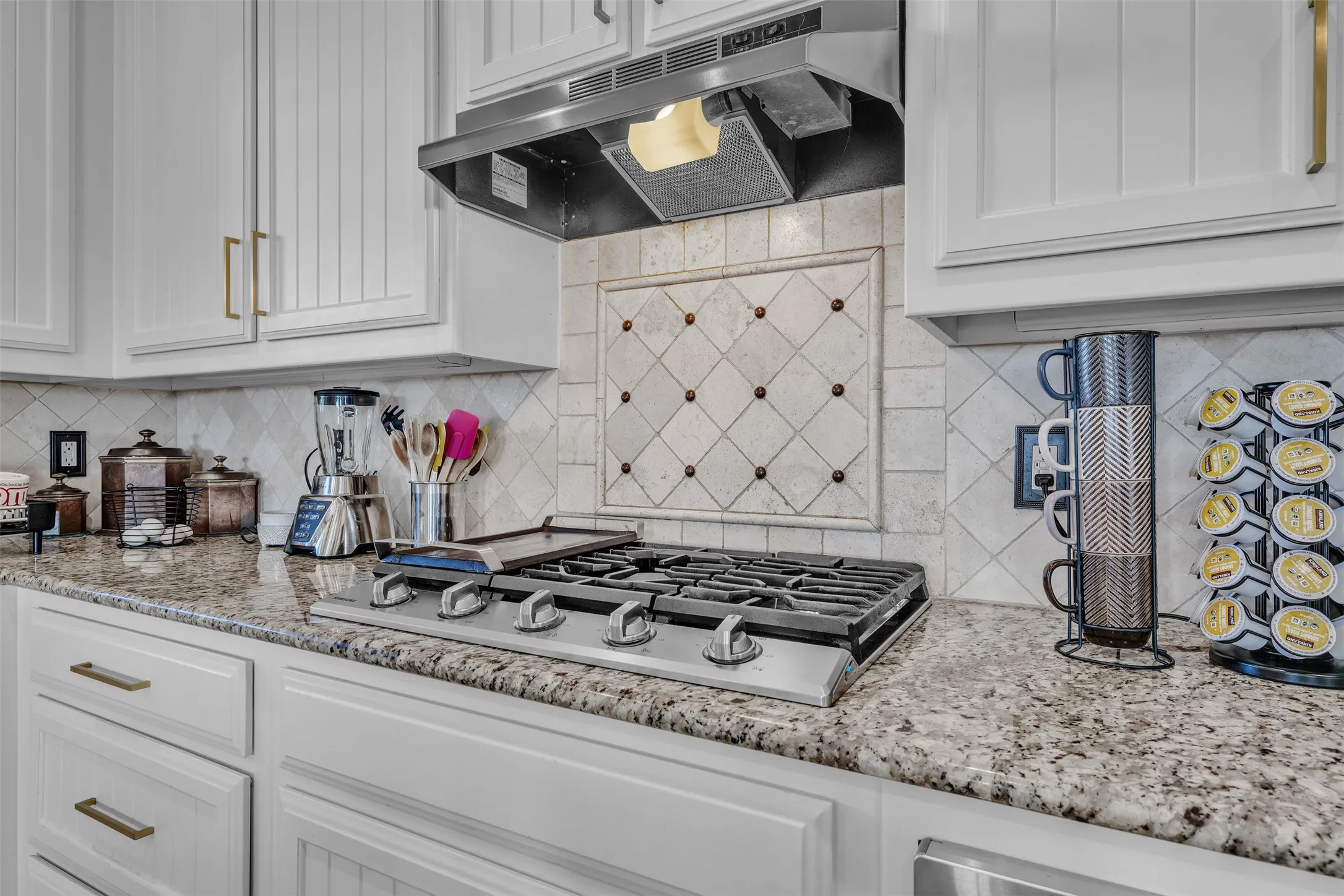 White shaker cabinetry with gold-tone hardware, stainless steel range hood, stone-finish countertops, tiled backsplash with decorative inset, and a built-in gas range