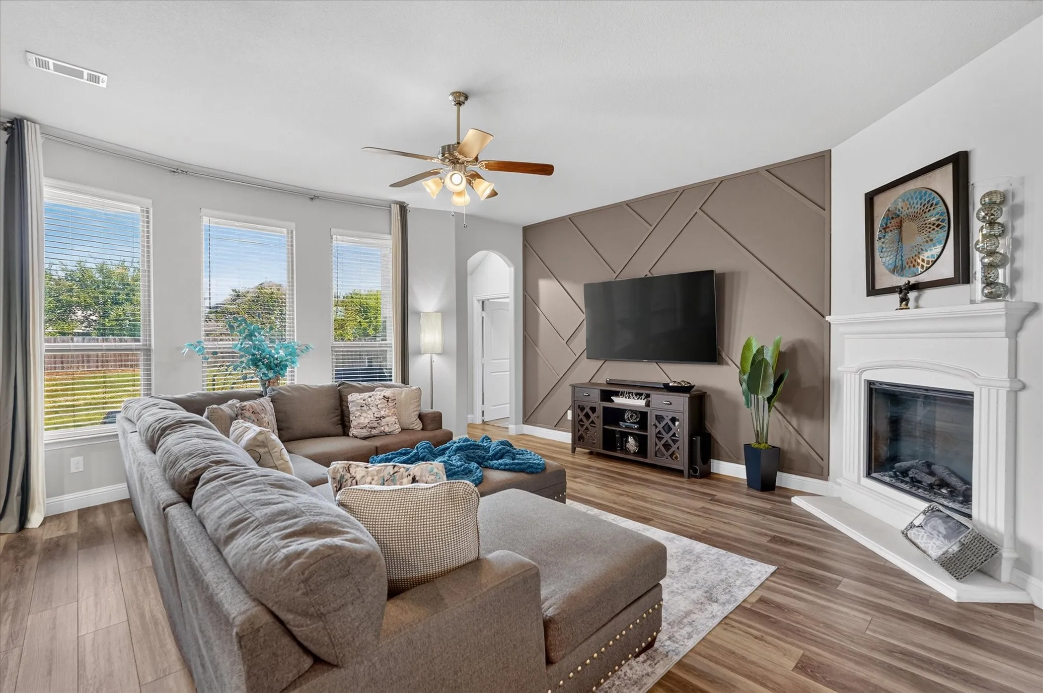 Wood-finish flooring, bay windows, ceiling fan with integrated lighting, decorative wall paneling, and a built-in fireplace