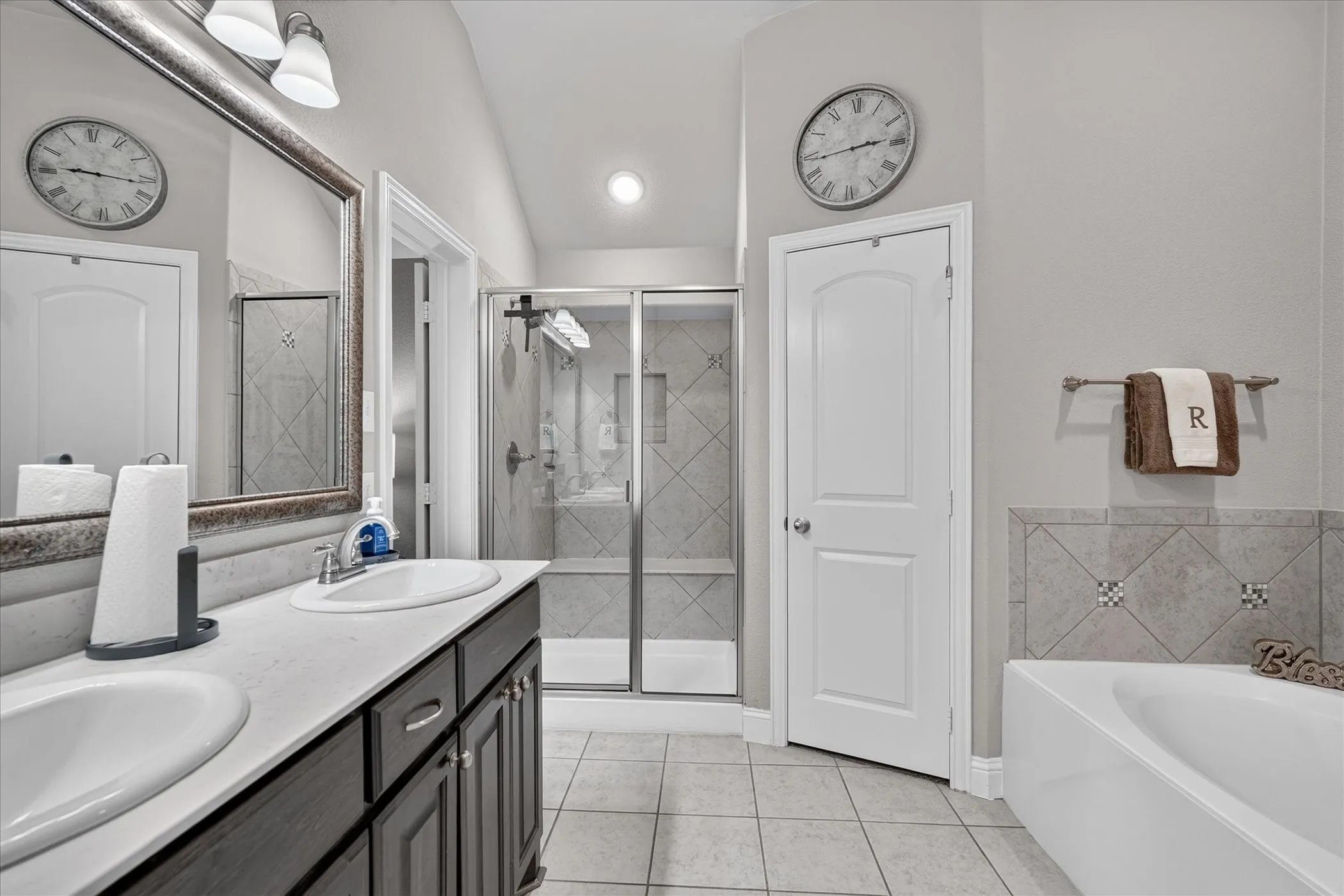 Dual vanity with integrated sinks and dark wood-finish cabinetry, frameless glass shower enclosure, tiled tub surround, and ceramic tile flooring