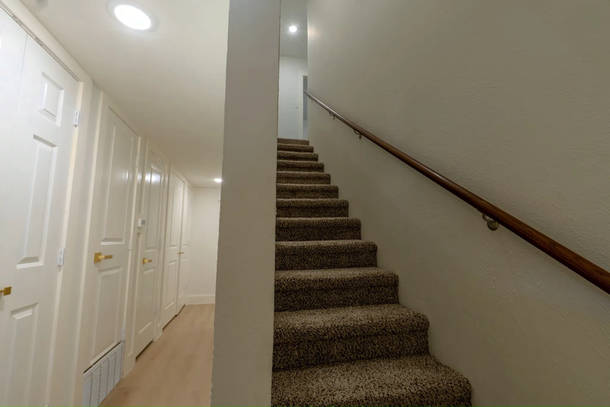 Carpeted staircase with a wood handrail, recessed lighting, and a hallway featuring multiple paneled doors with gold-finish hardware and wood-finish flooring