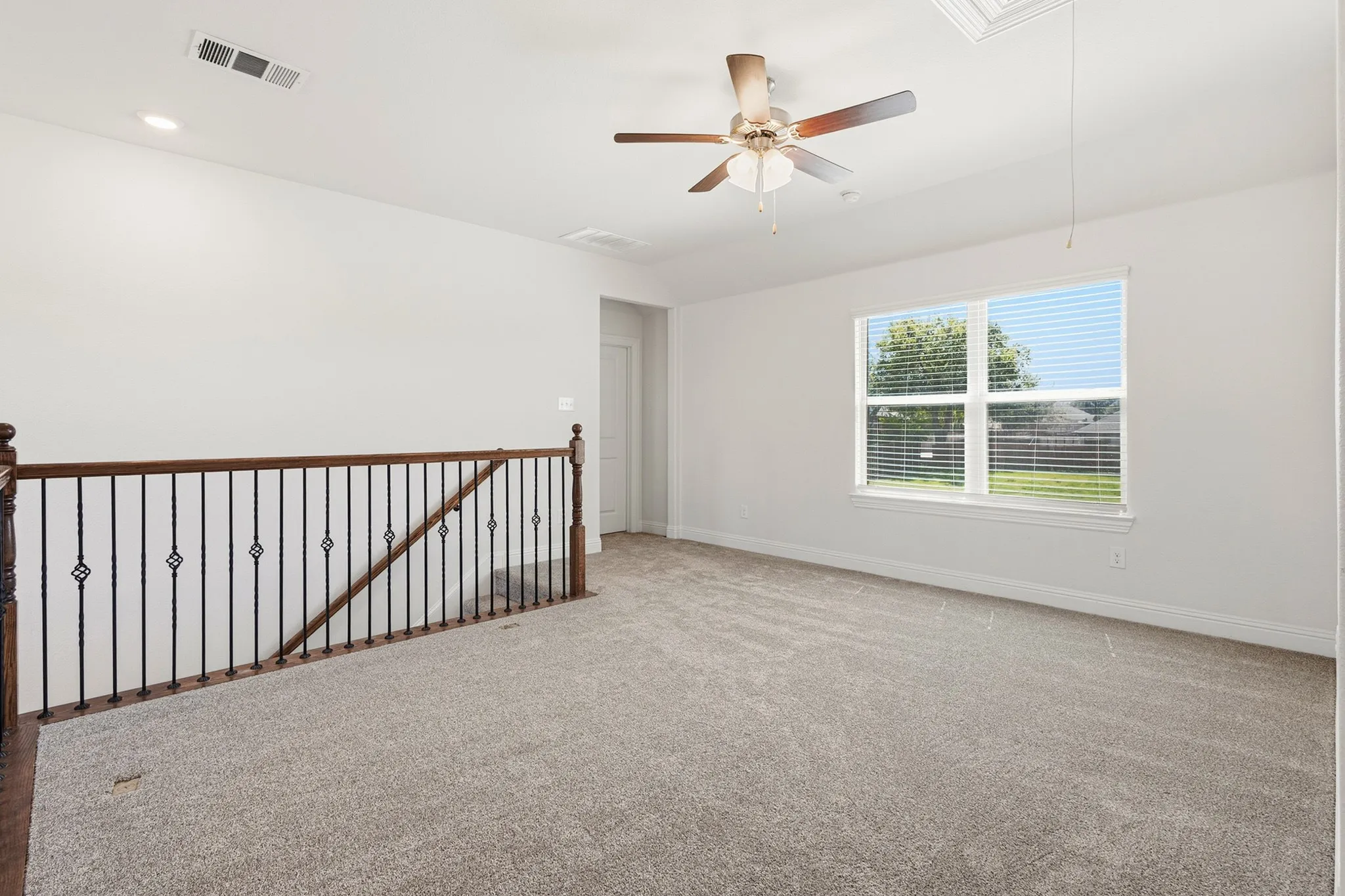 Carpeted flooring with a large window, ceiling fan, recessed lighting, and a wood-finish railing with iron balusters