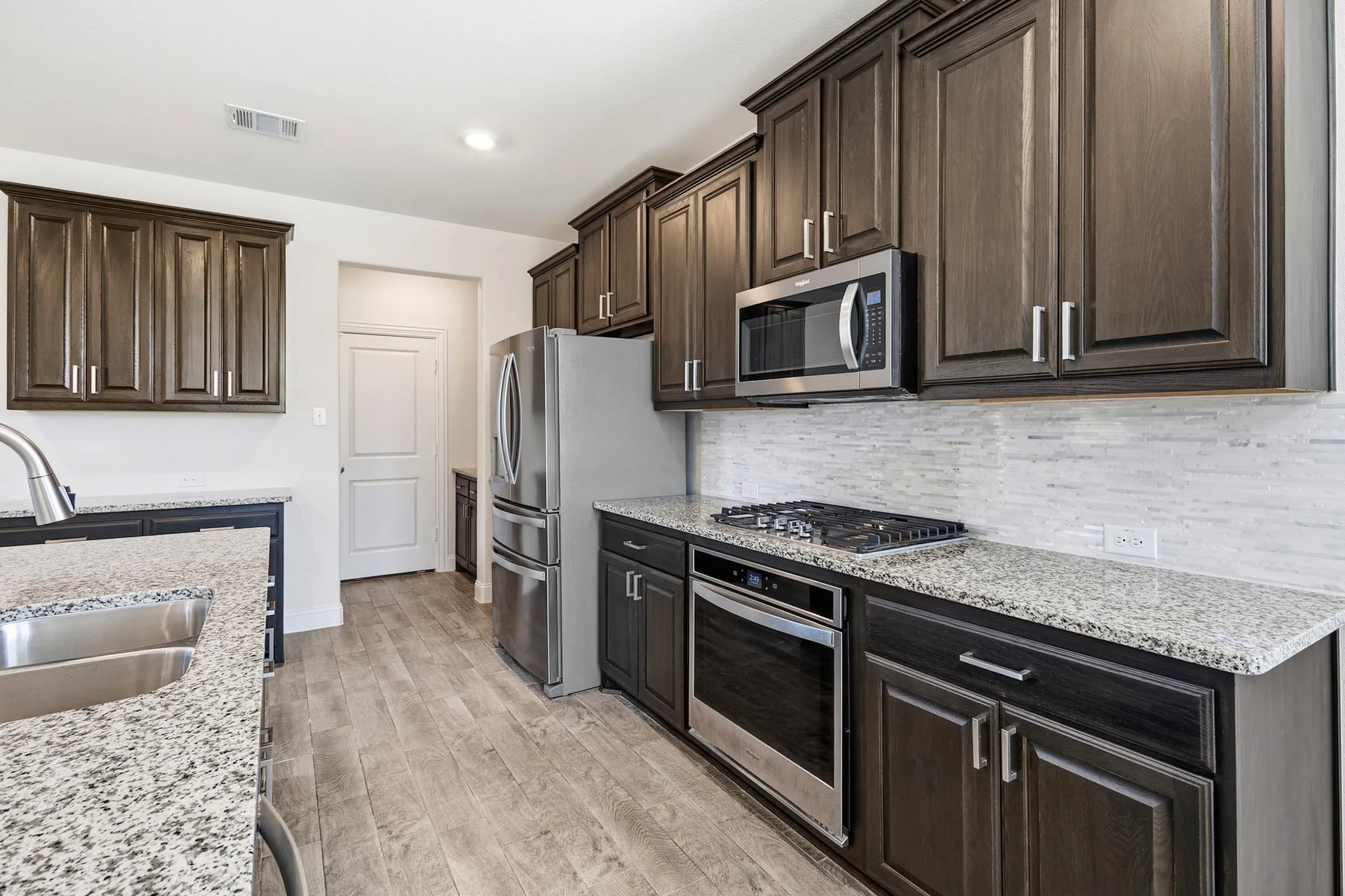 Kitchen featuring wood-finish flooring, dark wood cabinetry with stainless hardware, light-colored speckled countertops, a stainless steel sink with a gooseneck faucet, and a horizontally stacked tile backsplash