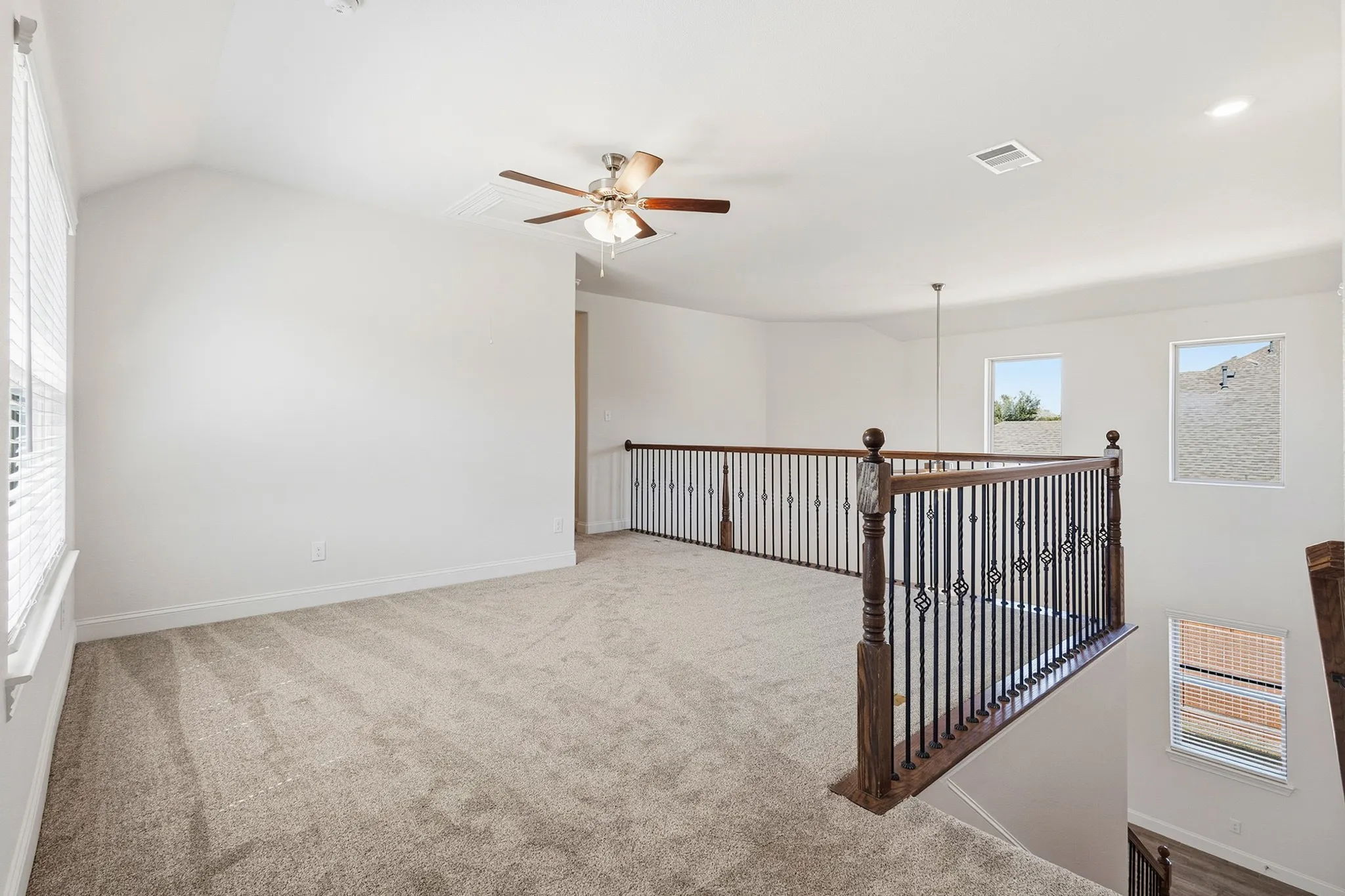 Carpeted loft area featuring a ceiling fan, multiple windows, and a wood and wrought iron railing