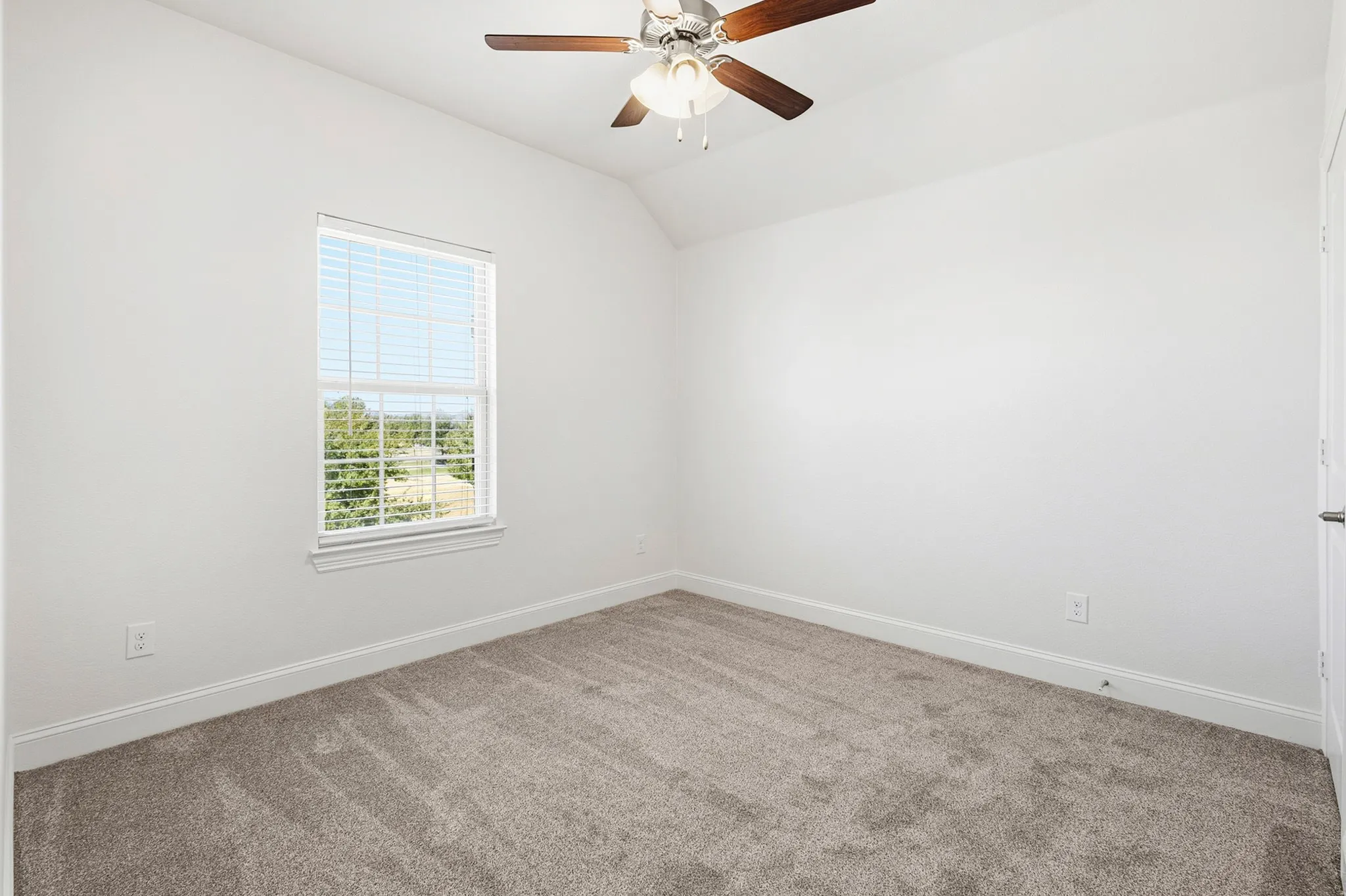 Light gray carpet, vaulted ceiling, white walls, window with blinds, and a ceiling fan with light fixture