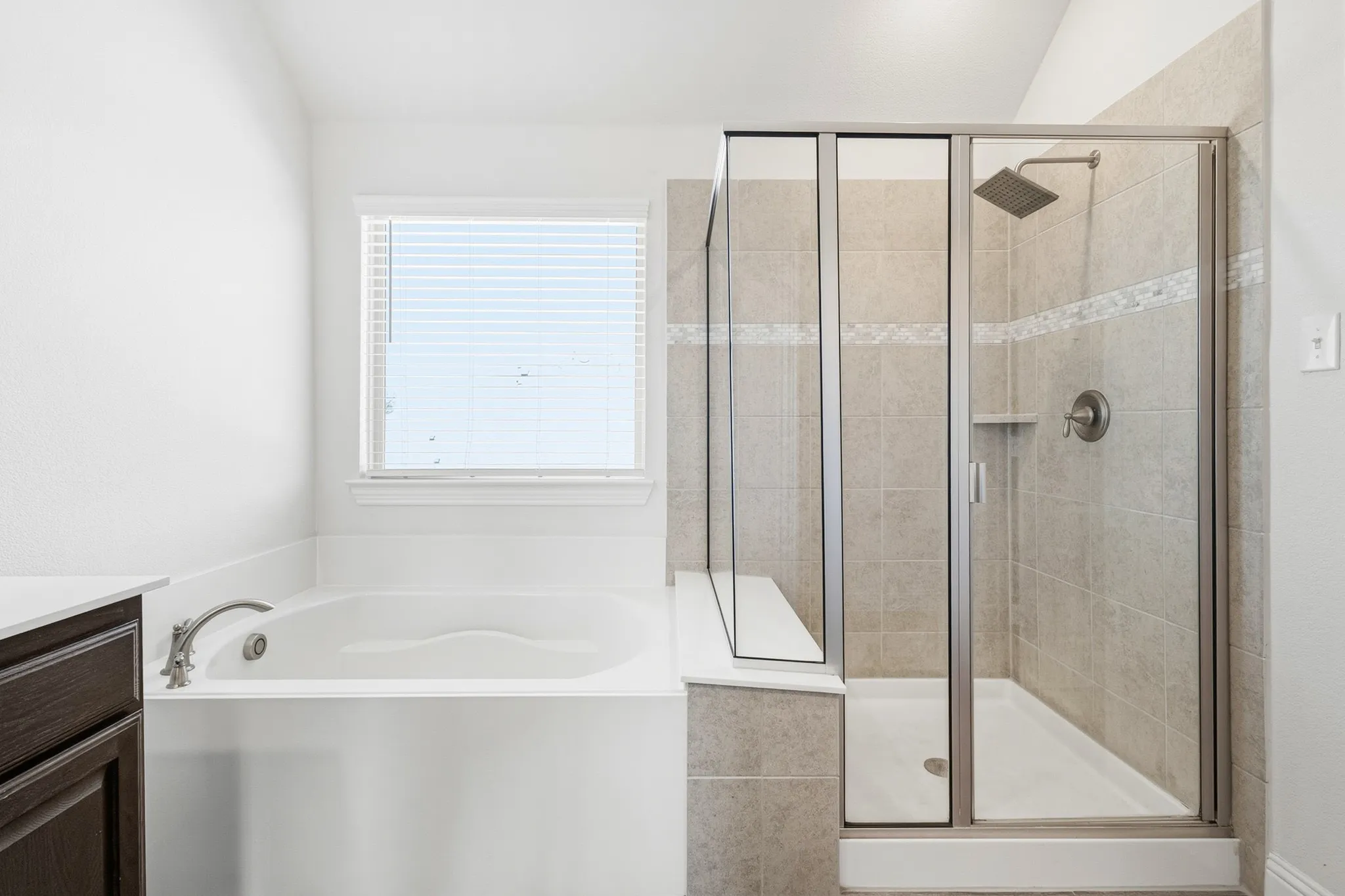 Built-in soaking tub, separate glass-enclosed shower, rain shower head, tiled shower surround with accent band, and dark wood-finish vanity