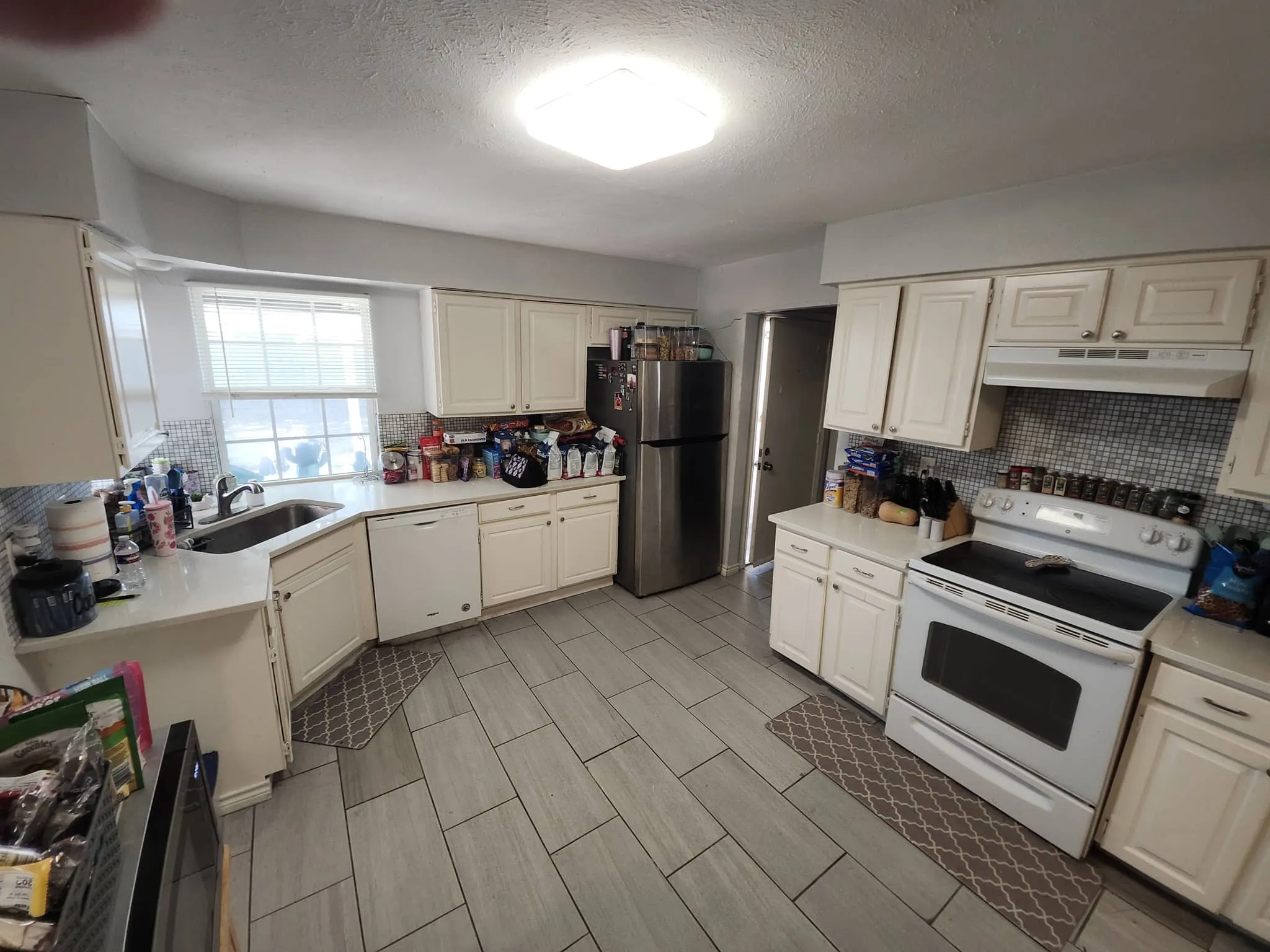 White cabinetry with brushed nickel hardware, L-shaped countertop, stainless steel sink with gooseneck faucet, light grey tile backsplash, and light grey wood-finish flooring