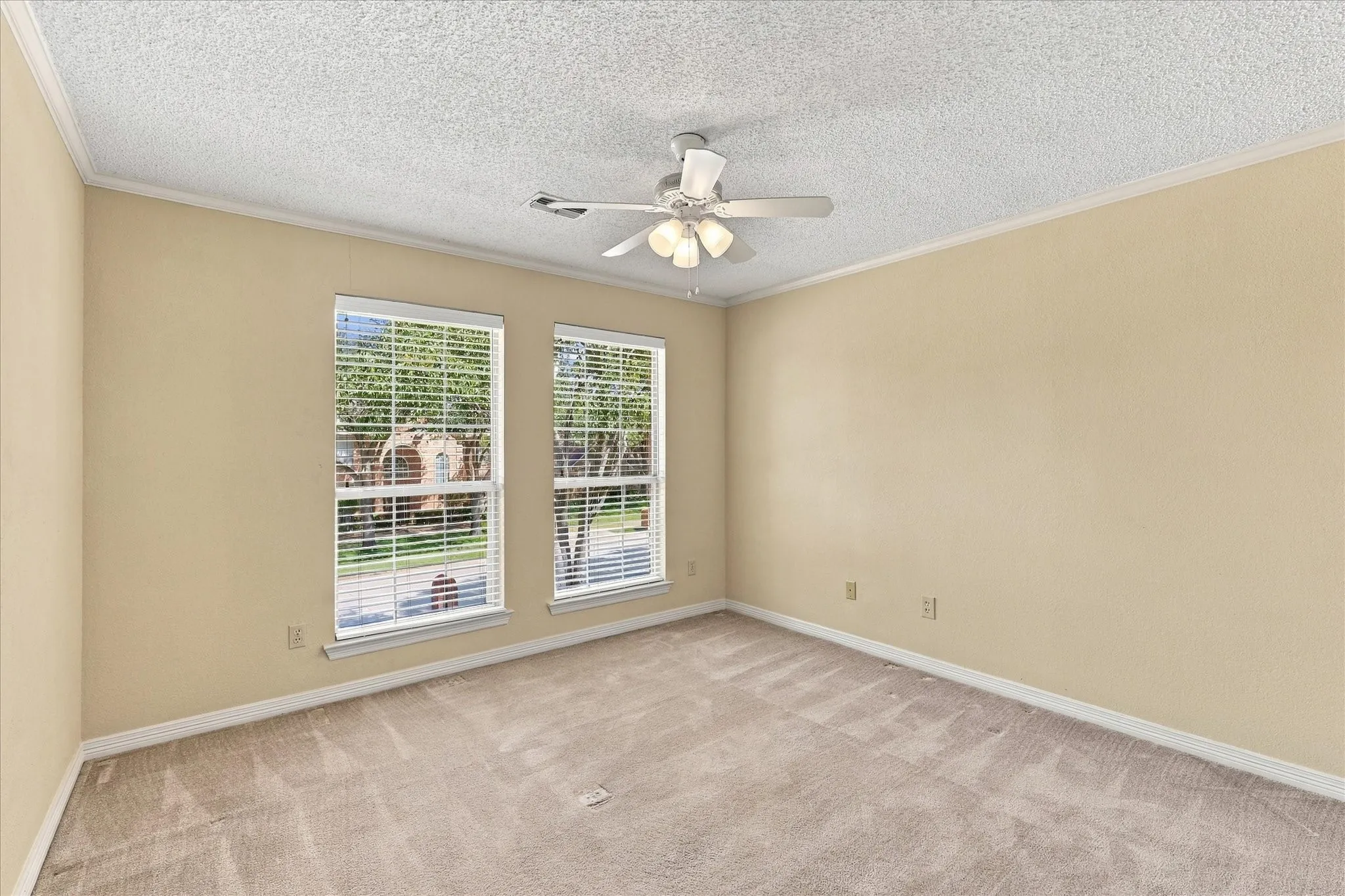 Neutral wall color, carpeted flooring, two windows with blinds, ceiling fan with light fixture, crown molding detail