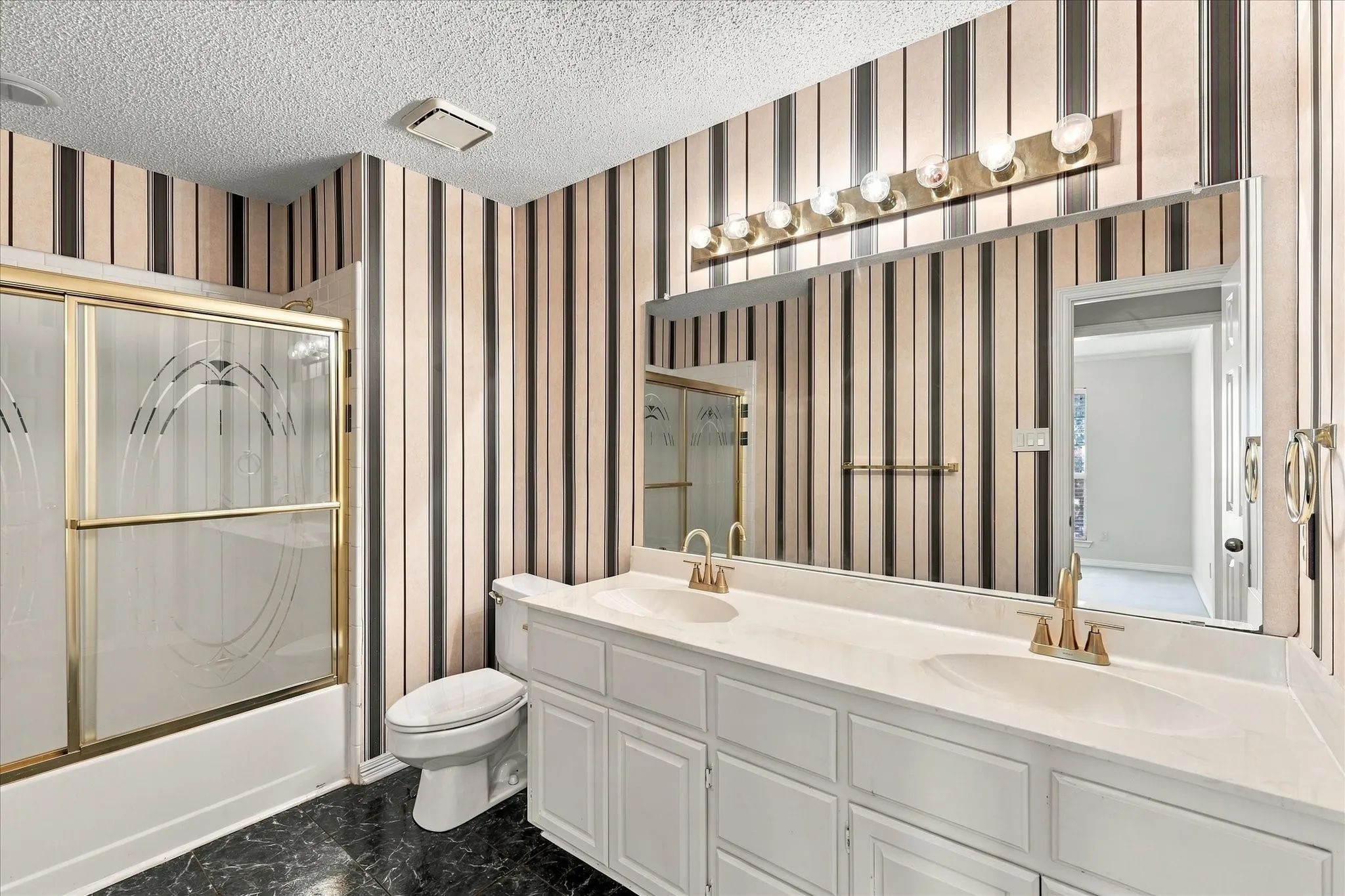Dual sink vanity with brass-finish fixtures, a large wall-mounted mirror, and a shower-over-tub with sliding glass doors