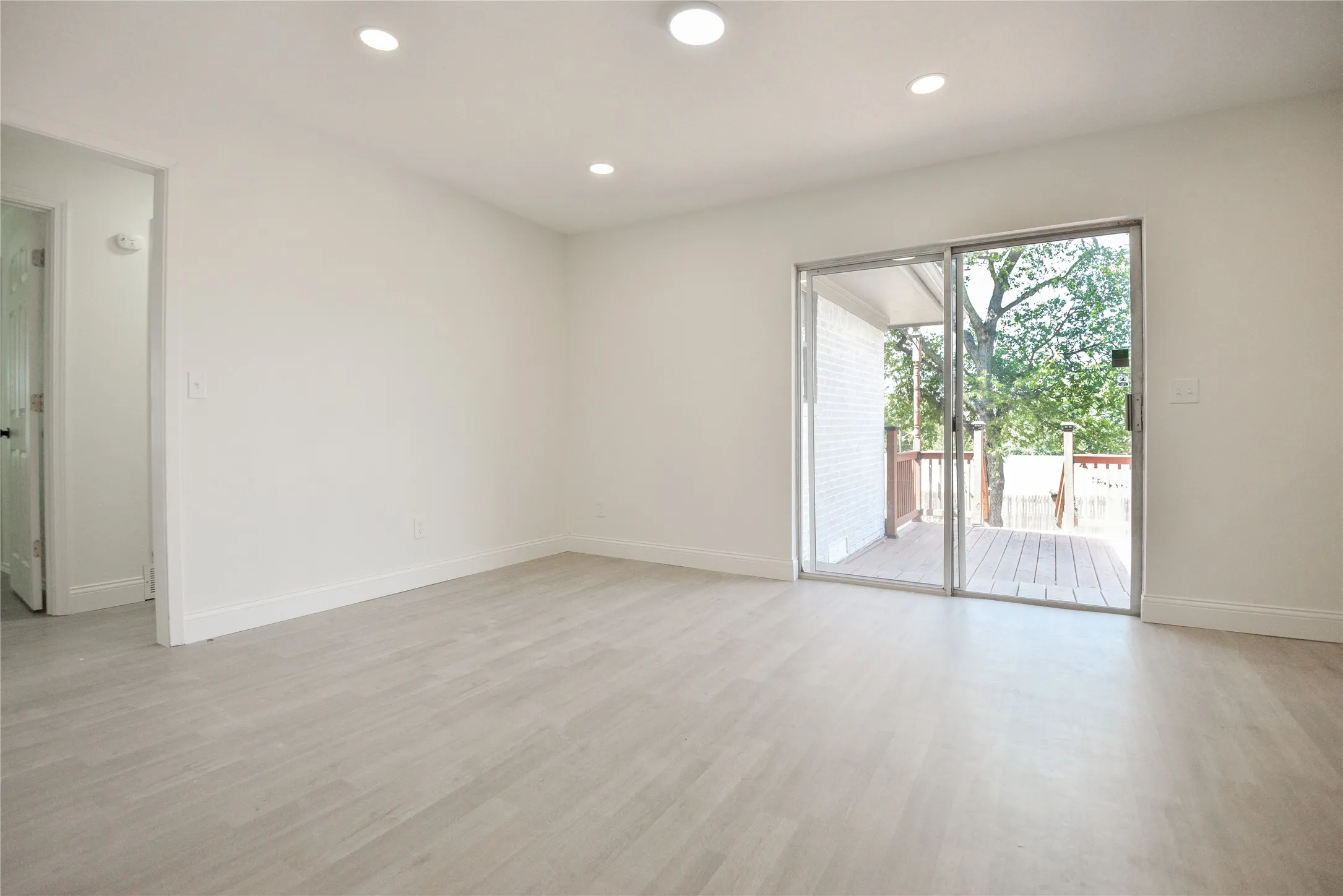 Recessed ceiling lighting, light wood-finish flooring, white painted walls, and a sliding glass door providing access to an outdoor deck with wood railings