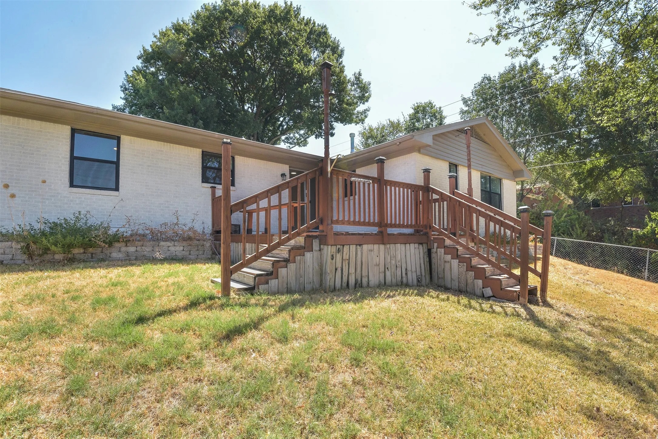 Wood deck with dual staircases and wood railing, white brick exterior, black-framed windows, and a retaining wall