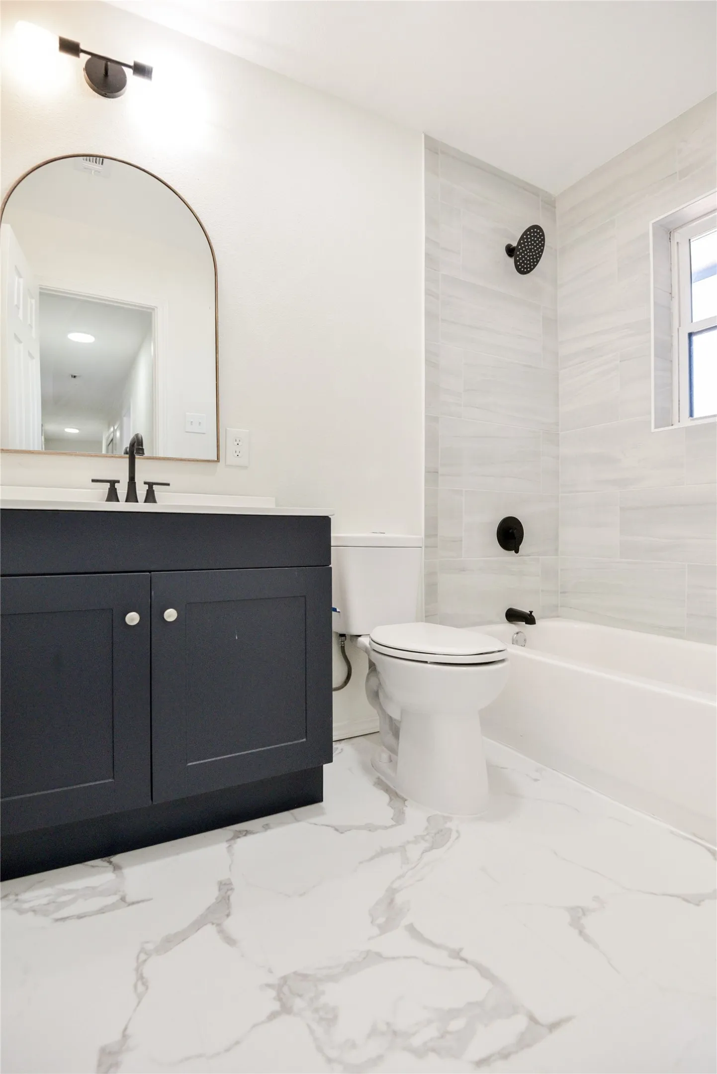 Modern bathroom featuring a dark navy vanity with polished chrome hardware, white marble-pattern porcelain tile flooring, white subway tile shower surround, arched mirror with integrated lighting, and matte black fixtures including a rainfall showerhead and wall-mounted faucet