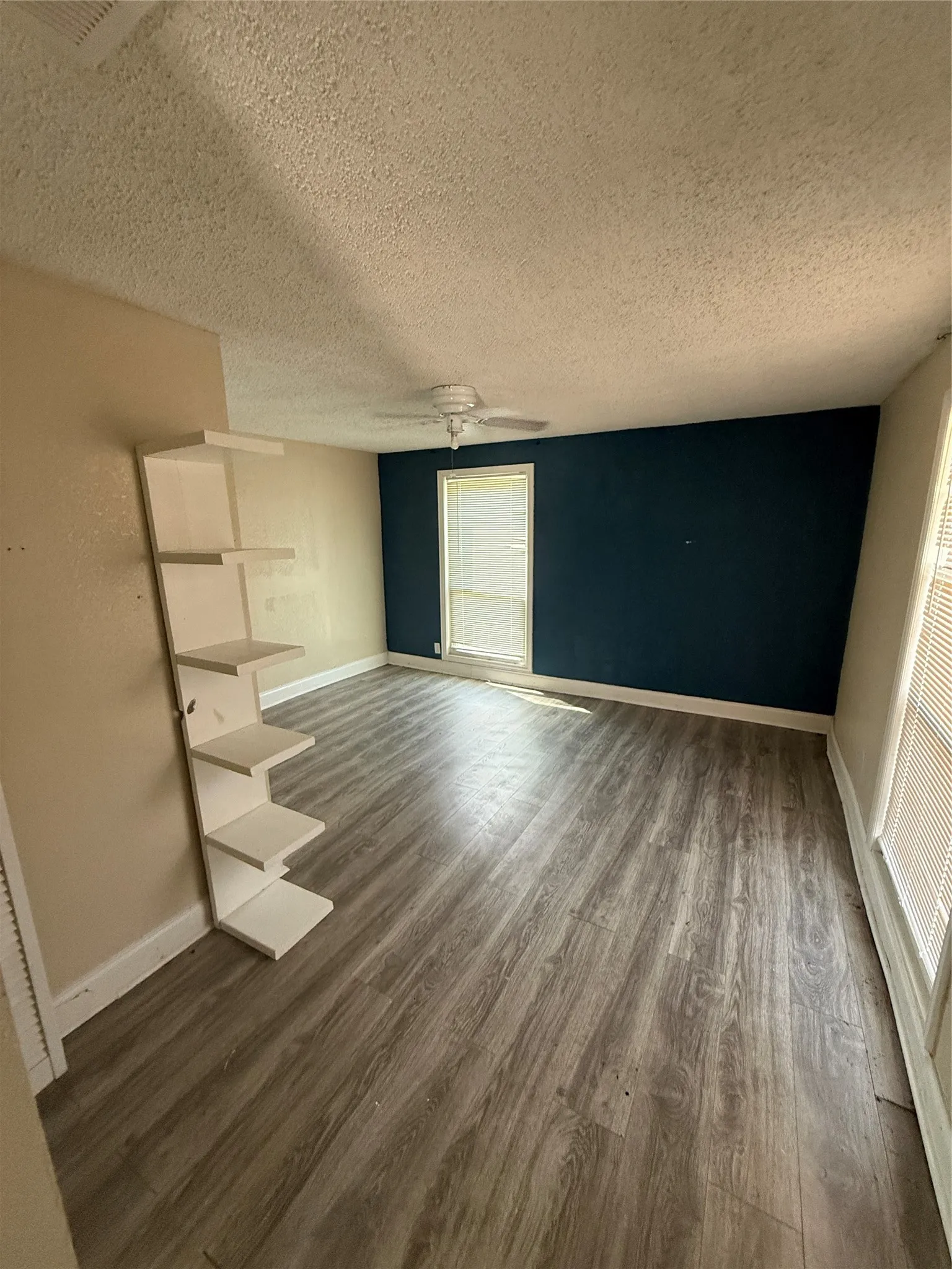 Wood-finish flooring, built-in shelving, ceiling fan, window with blinds, and a contrasting accent wall