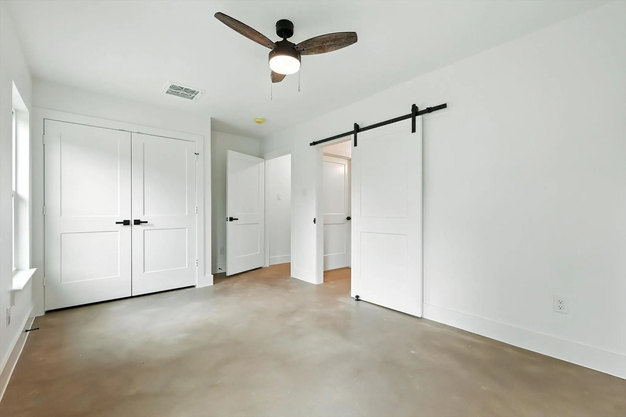 Smooth concrete flooring, white walls, ceiling fan with integrated lighting, double panel doors with black hardware, and a white barn door on a black track