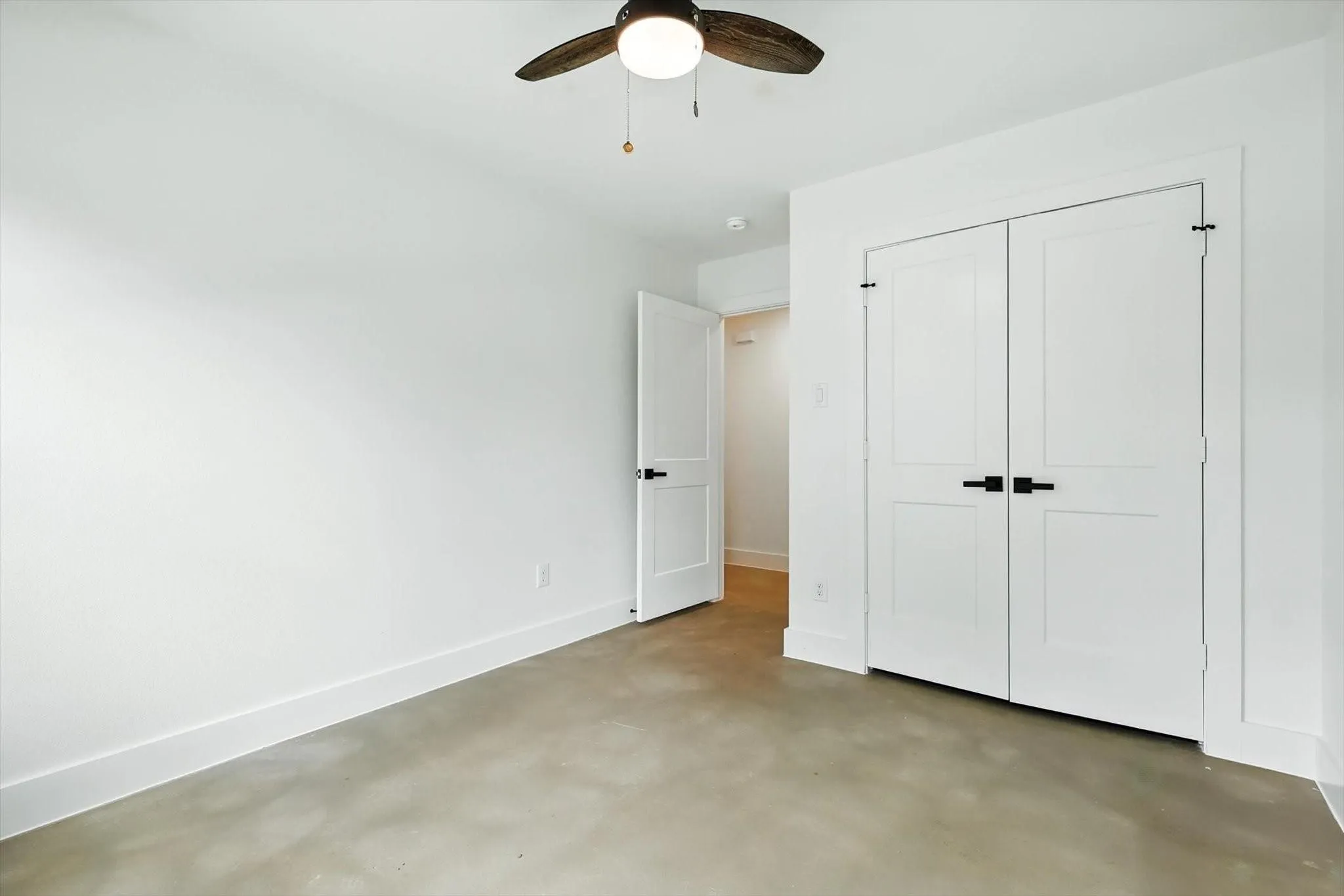 Ceiling fan with light fixture, double-door closet with matte black hardware, white walls, and concrete flooring