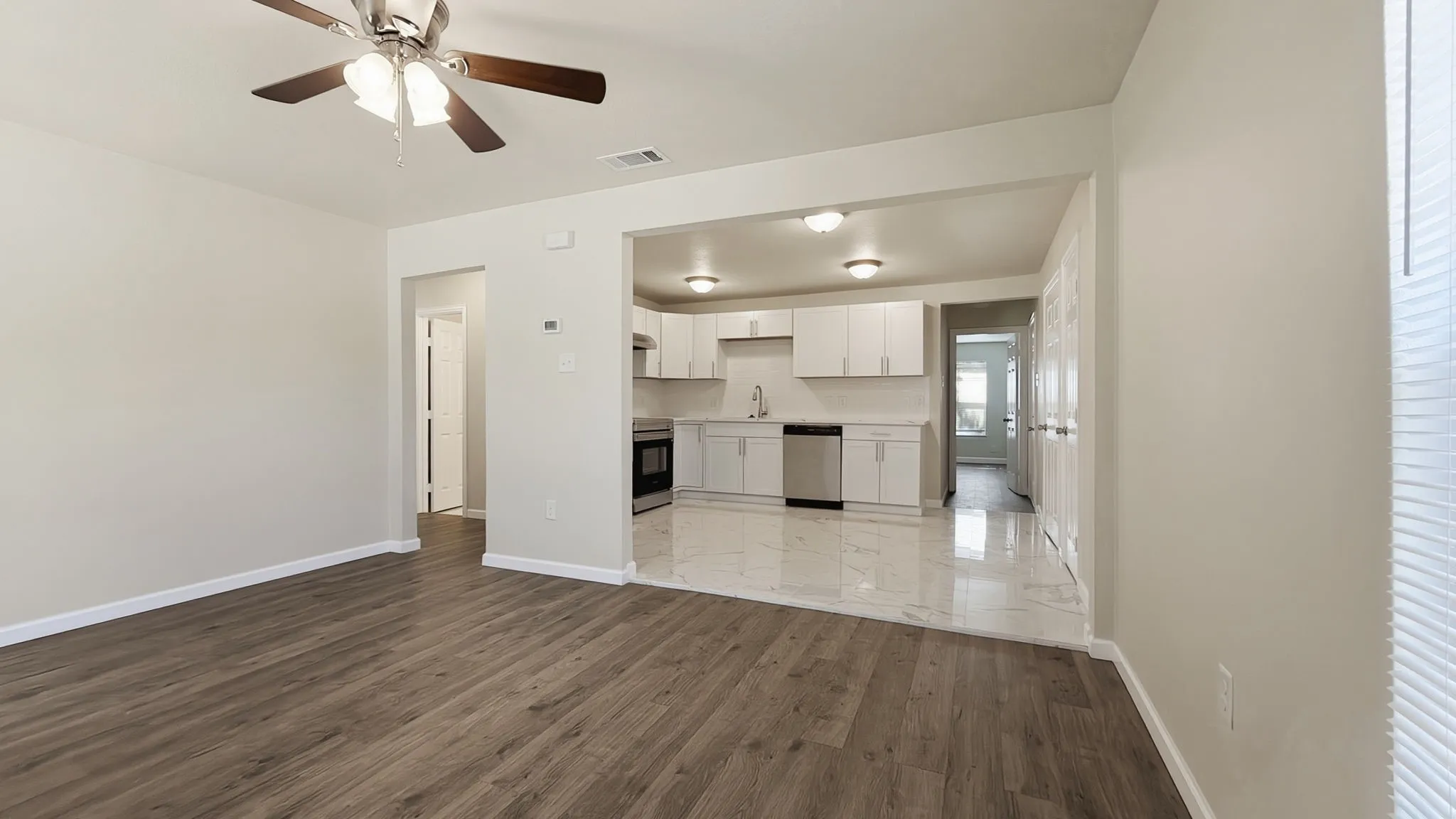 Wood-finish flooring with a ceiling fan, leading into a kitchen with white cabinetry, stainless steel appliances, and polished tile flooring