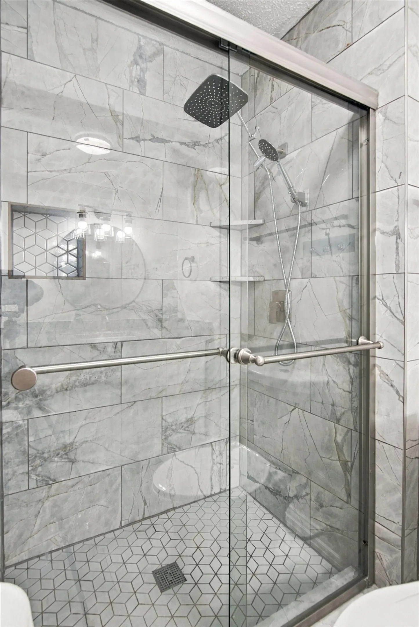Glass-enclosed shower with a rainfall showerhead, handheld sprayer, geometric tile flooring, and large format wall tiling