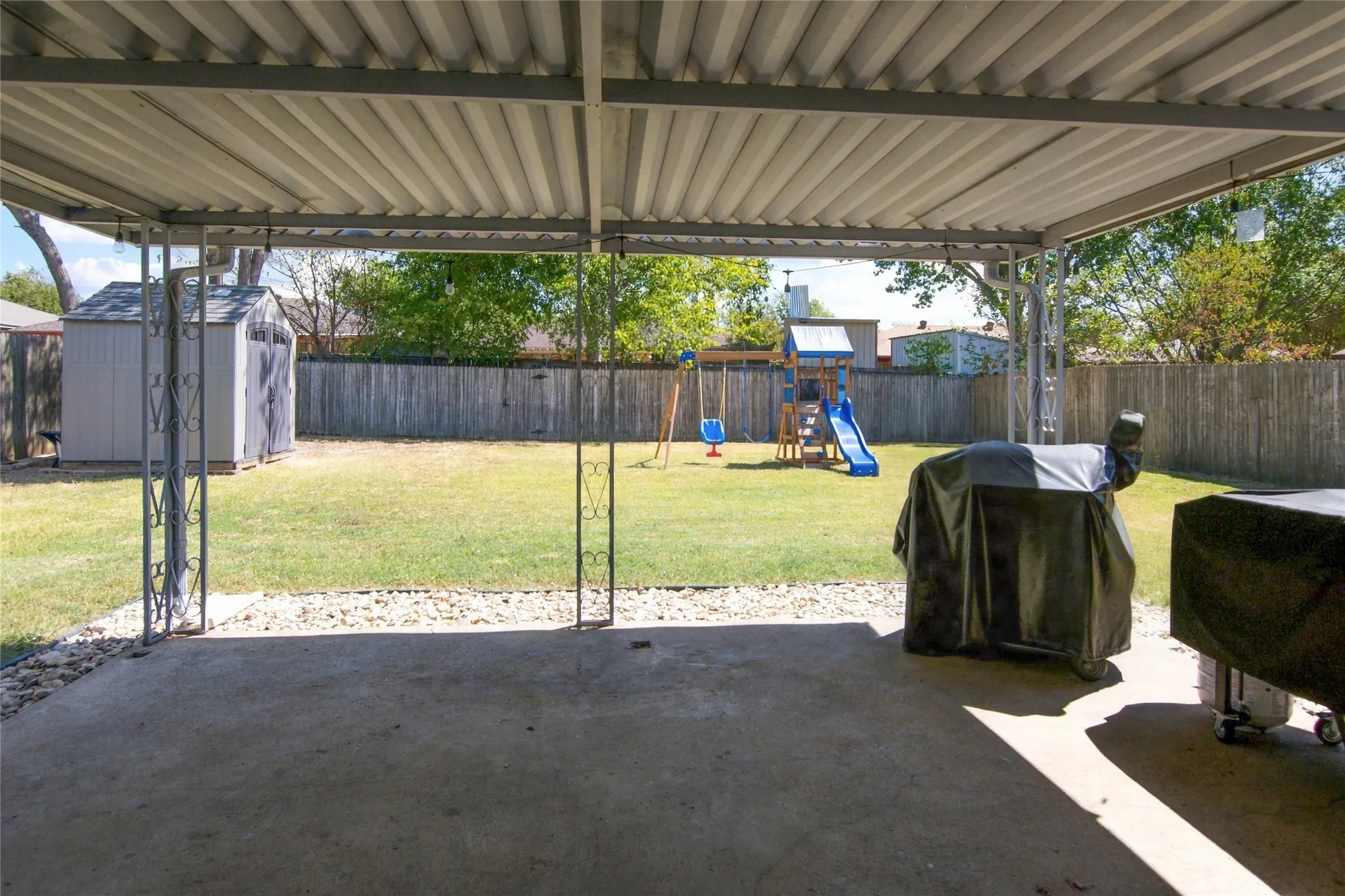 Covered concrete patio with decorative metal supports, overlooking a grassy yard with a privacy fence and a storage shed
