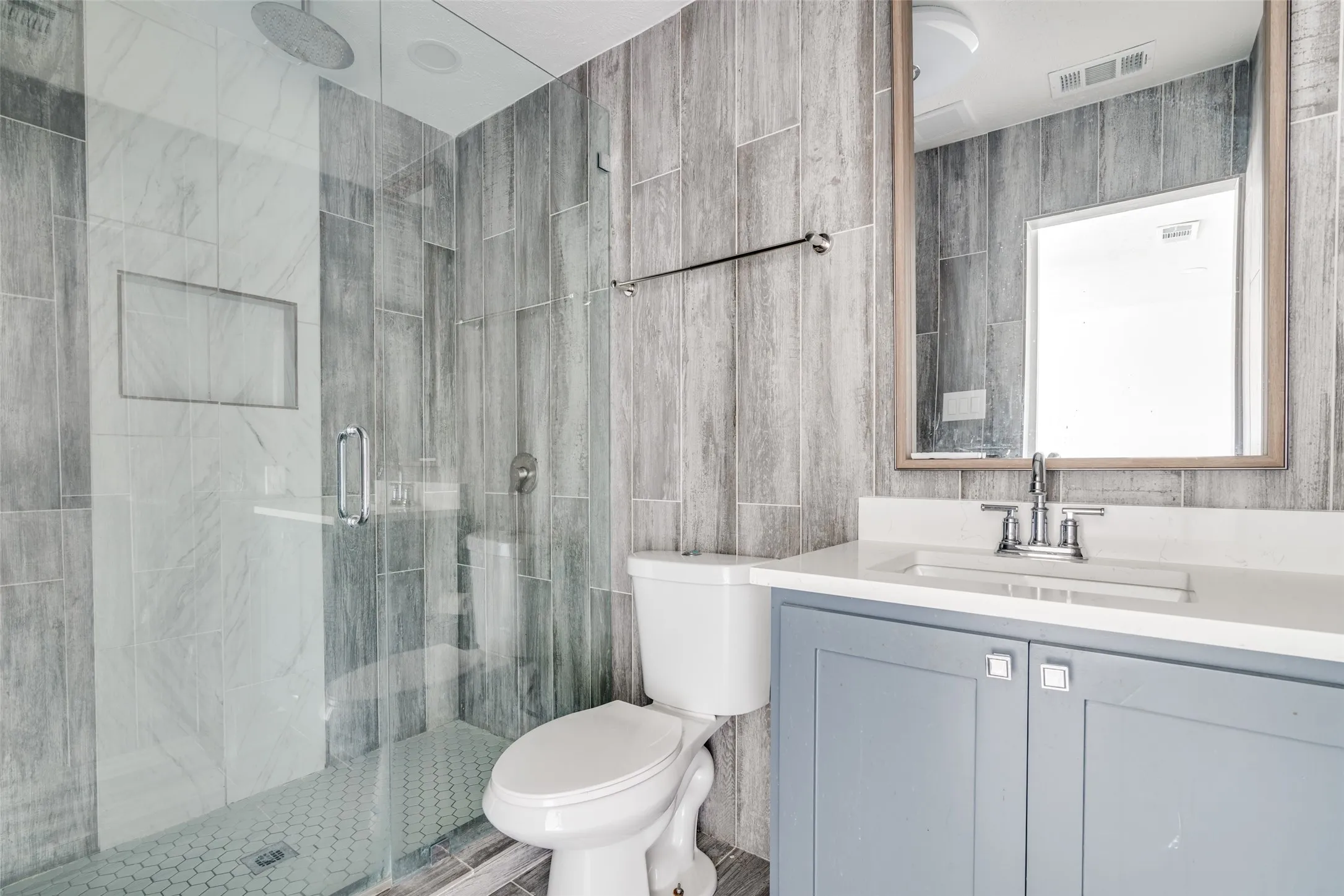 Glass-enclosed shower with a rainfall showerhead and tiled niche, wood-finish wall tile, white vanity countertop, undermount sink, and a framed mirror