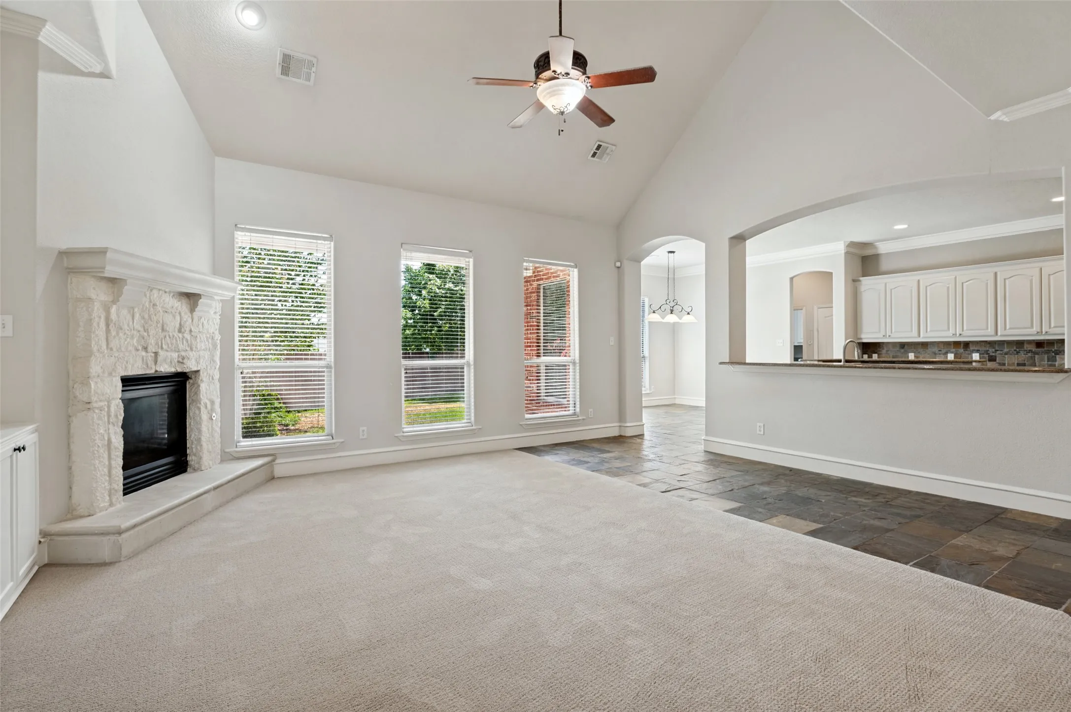 Vaulted ceilings, a stone-clad fireplace, and a ceiling fan enhance the living area, while a breakfast bar with white cabinetry and a tile backsplash separates the kitchen space