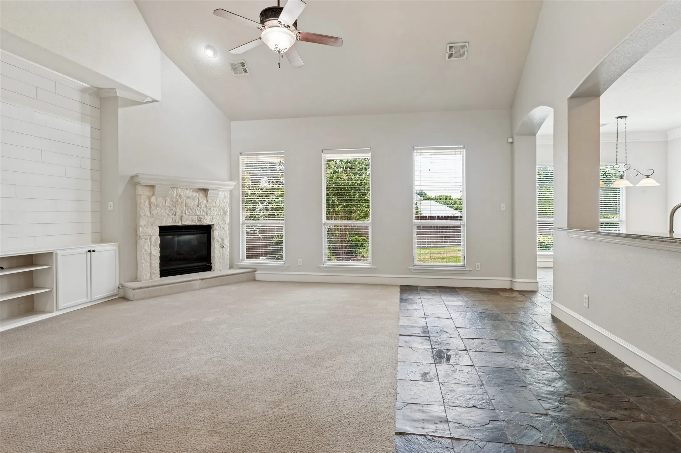 Vaulted ceiling with a ceiling fan, stone-clad fireplace, built-in cabinetry, large windows, and a mix of carpet and stone tile flooring