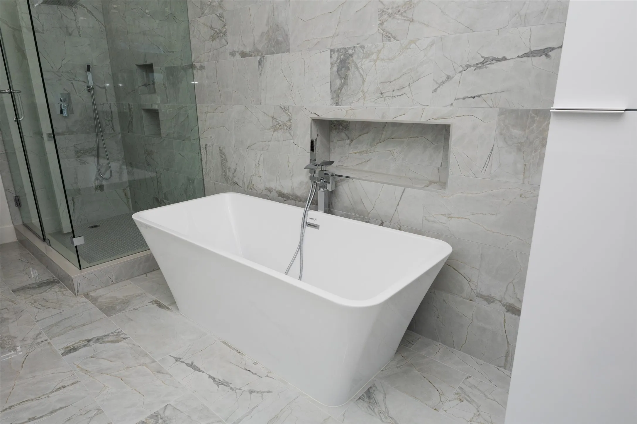 Freestanding soaking tub with floor-mounted faucet, glass-enclosed shower, large format wall and floor tile, and integrated shower niches