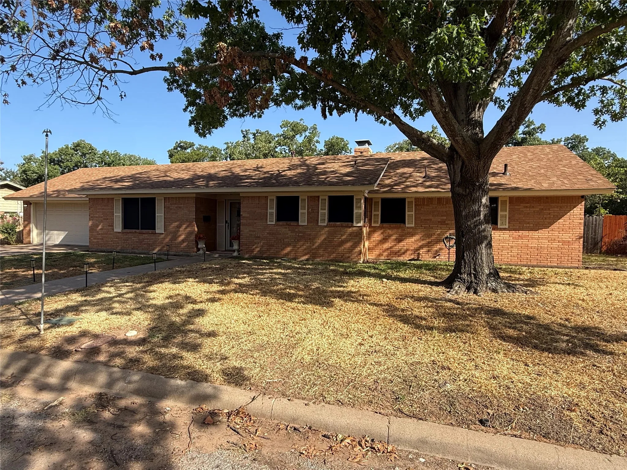 Brick exterior with a brown shingle roof, featuring white window shutters and a mature tree