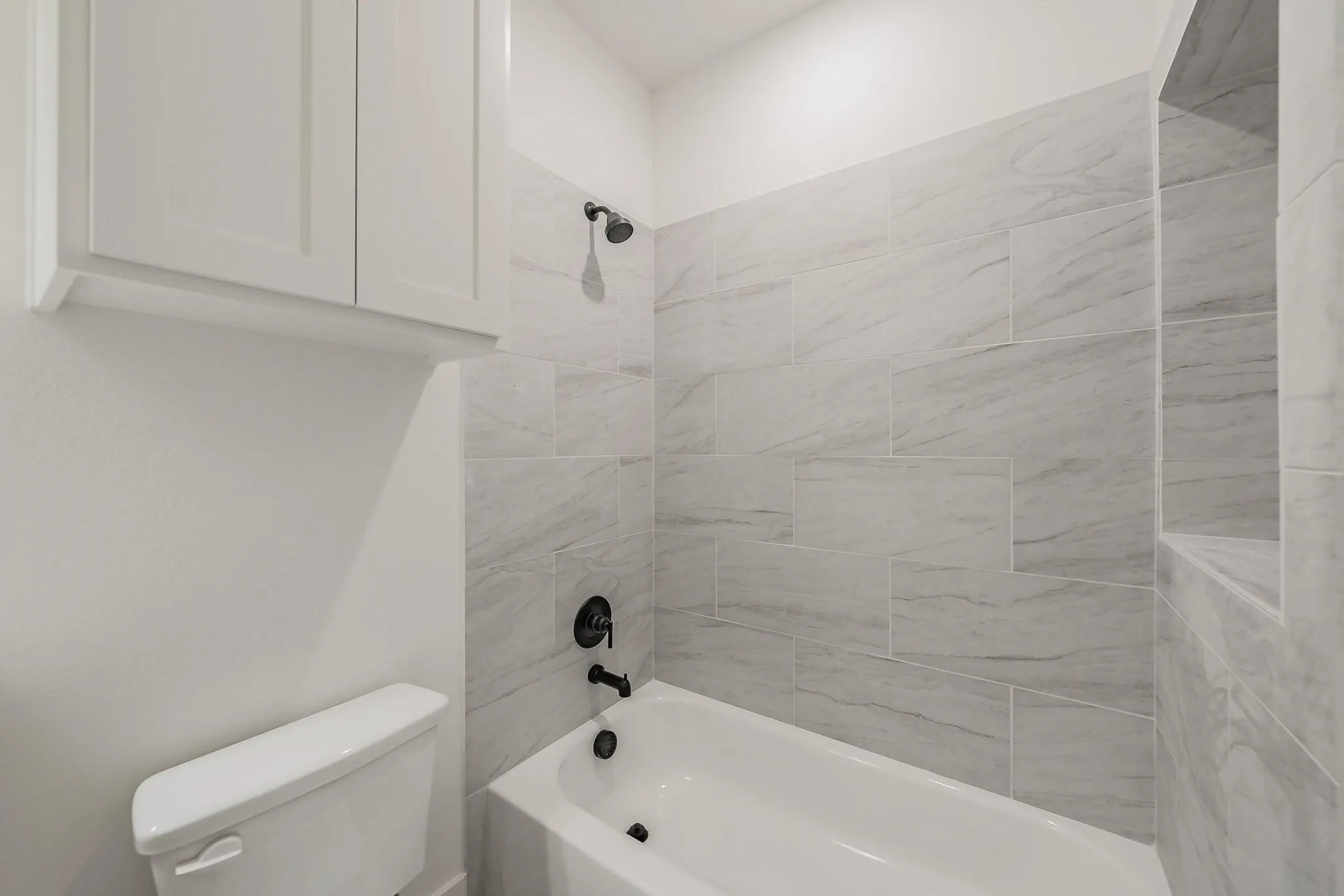 Bathtub with matte black fixtures, white subway tile surround, recessed shower niche, and white wall-mounted cabinetry