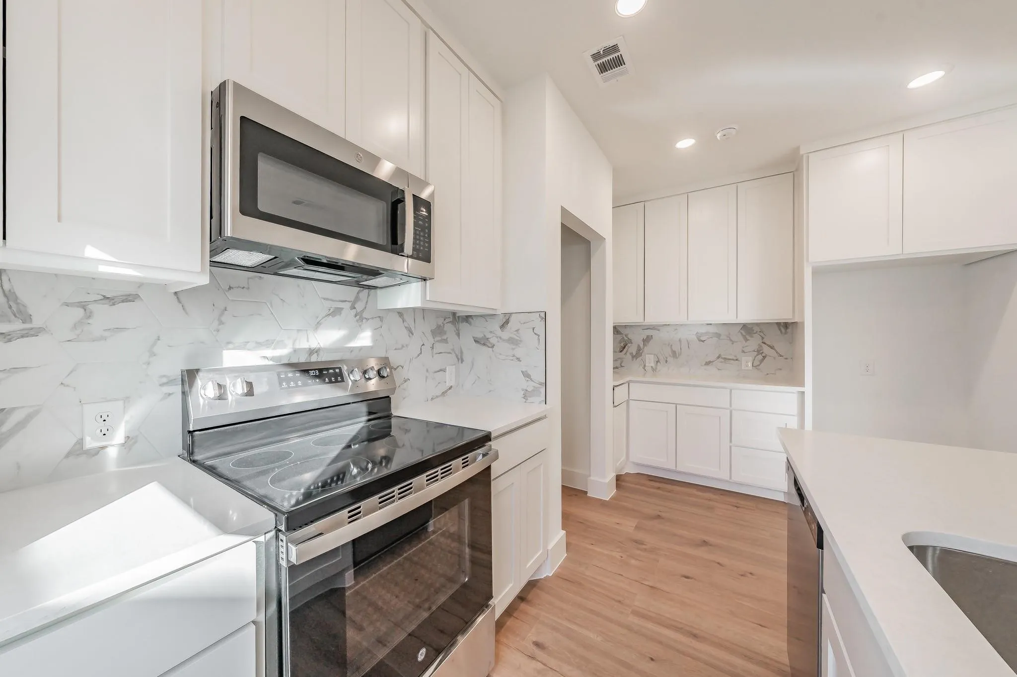 White shaker cabinetry, stainless steel appliances, light-toned wood-finish flooring, recessed lighting, and a white countertop with a gray-veined backsplash