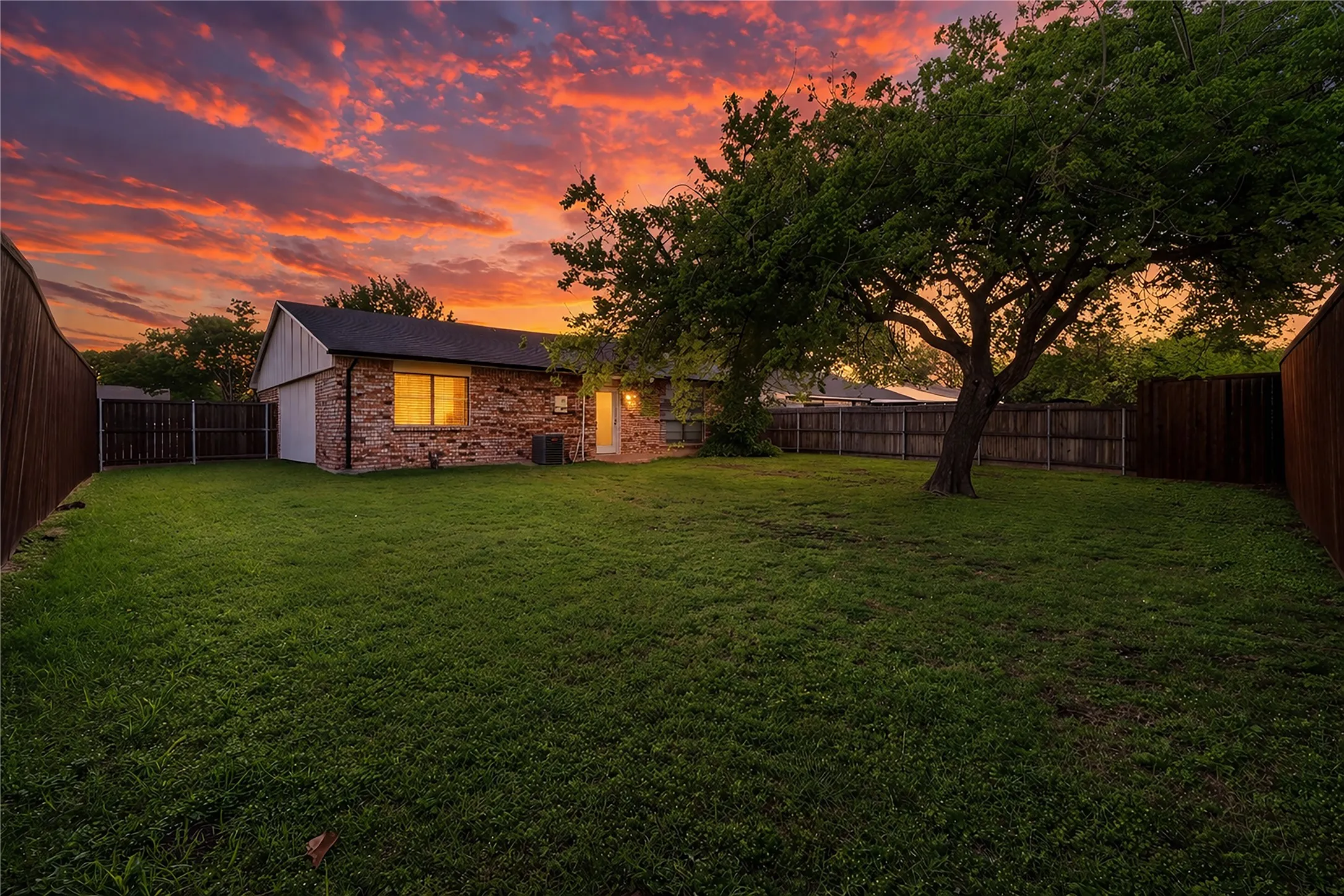 Expansive green lawn with mature tree, brick exterior, and wood fencing