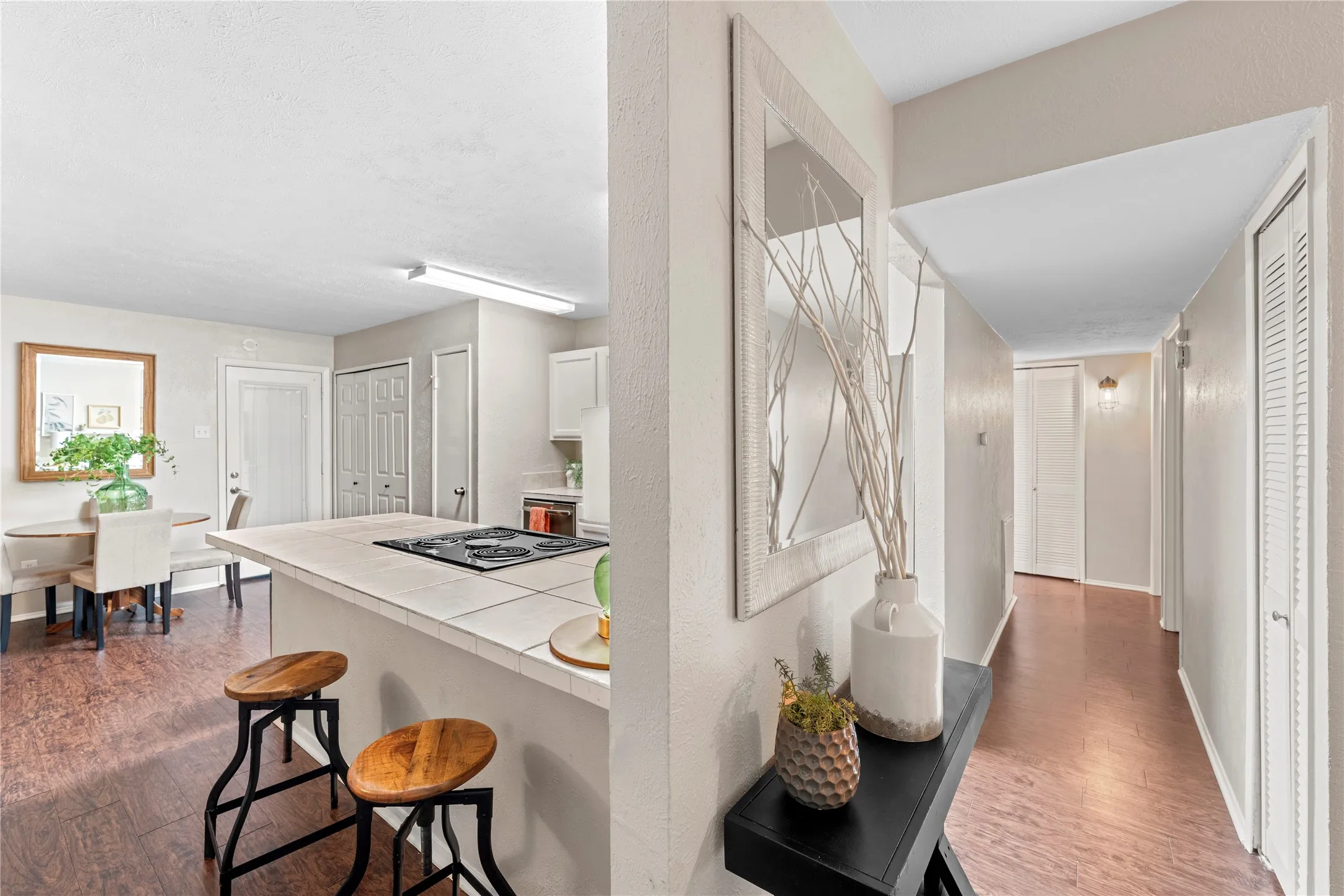 Wood-finish flooring throughout, kitchen island with built-in cooktop, white kitchen cabinetry, and a hallway with wall-mounted lighting