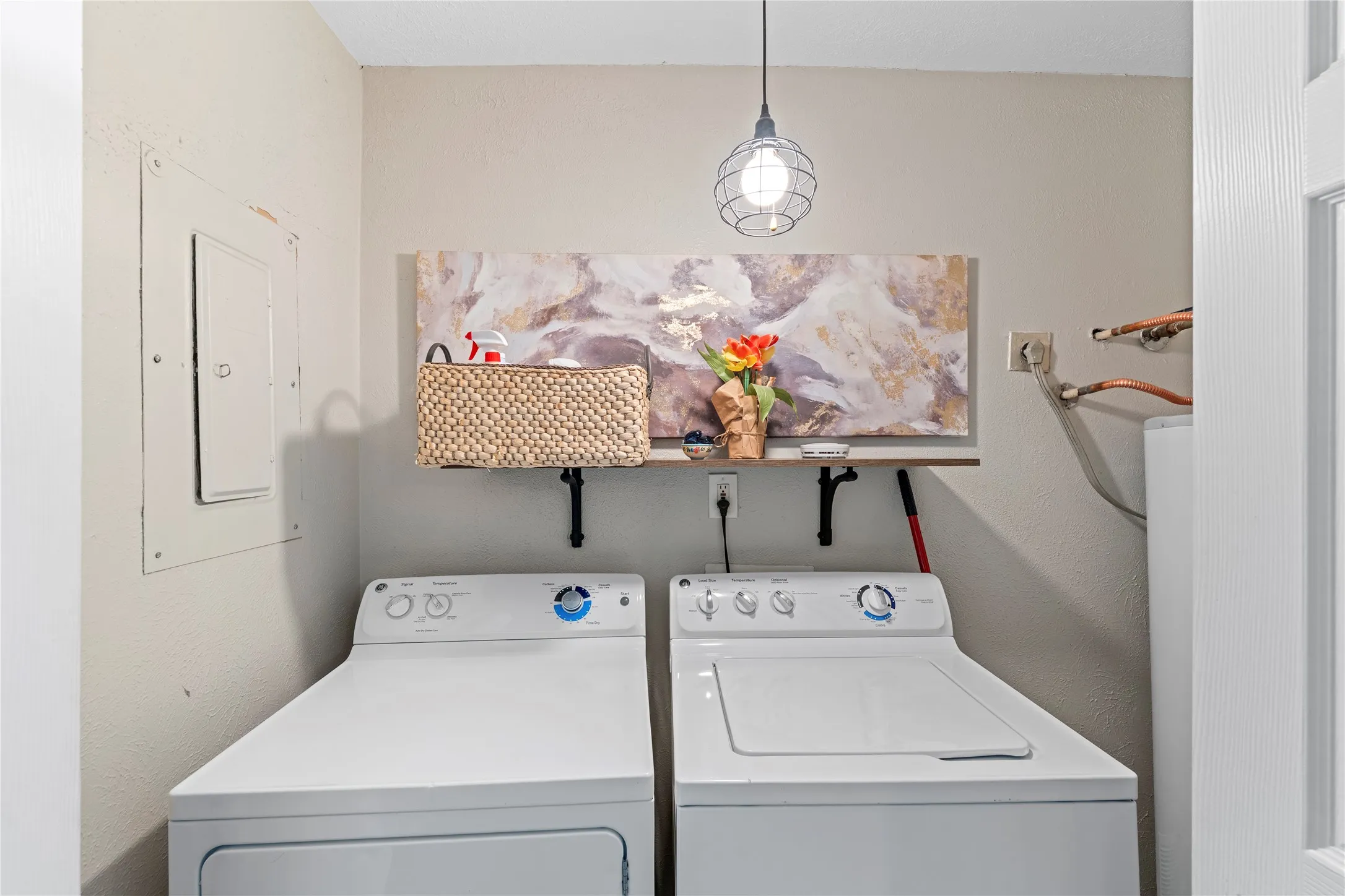 Utility area with two full-sized appliances, a suspended wire-frame light fixture, a floating wood-finish shelf, and wall-mounted electrical panel