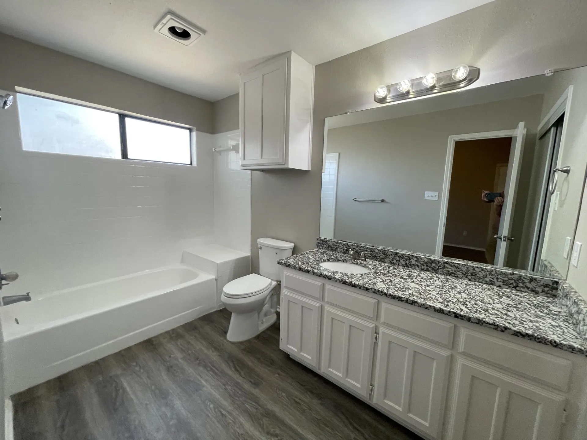 Dual-sink vanity with a light-toned countertop, wood-finish flooring, built-in white cabinetry, and a bathtub with white tile surround