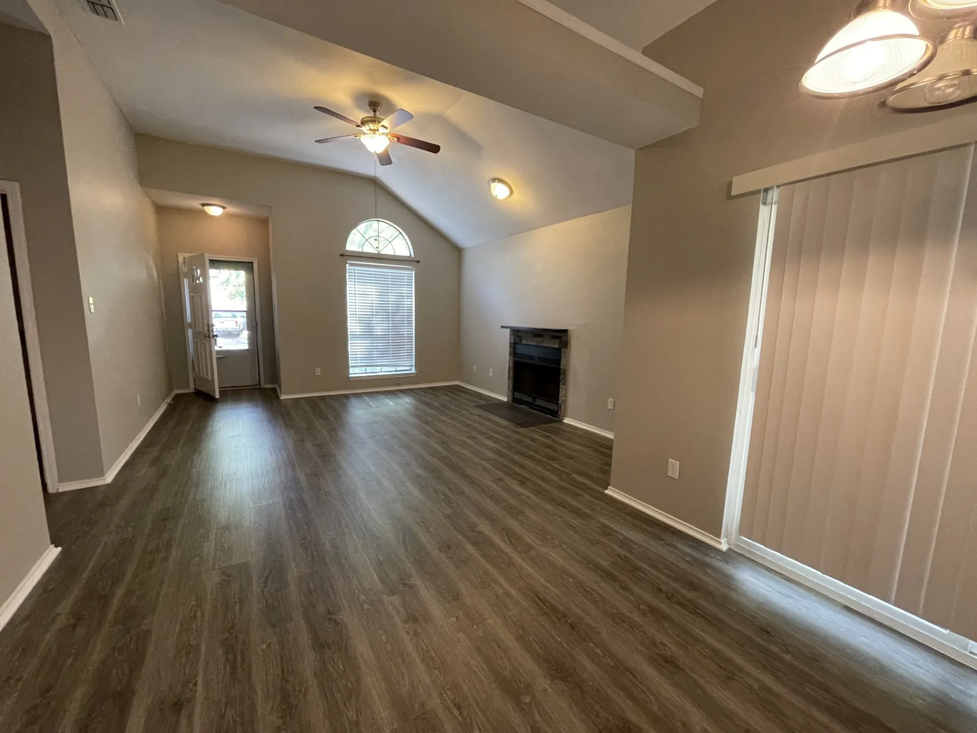 Vaulted ceiling, wood-finish flooring, ceiling fan, fireplace with dark stone surround, and vertical blinds