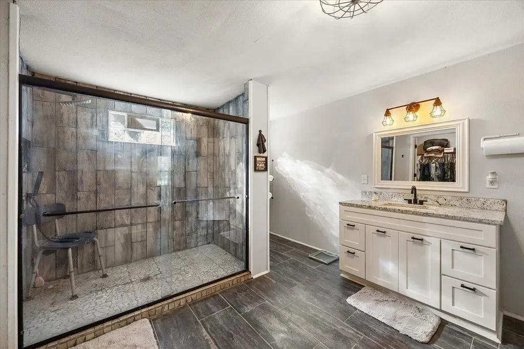 Primary bath alternate angle highlighting vanity mirror lighting, wood-look flooring, and tiled shower stall.