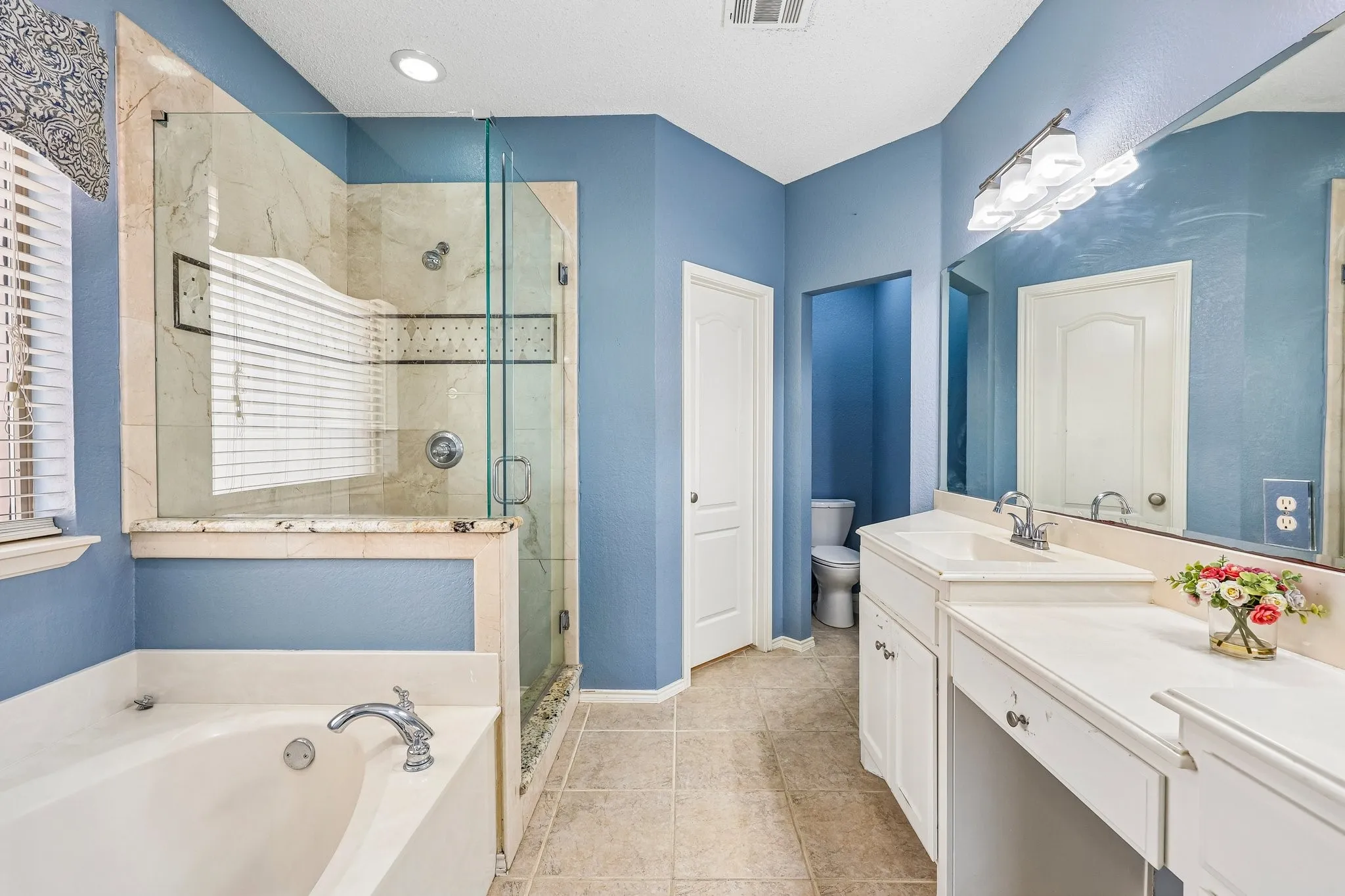 Bathroom featuring a glass-enclosed shower with stone-look tile, a separate soaking tub with deck-mounted faucet, and a dual vanity with integrated sinks