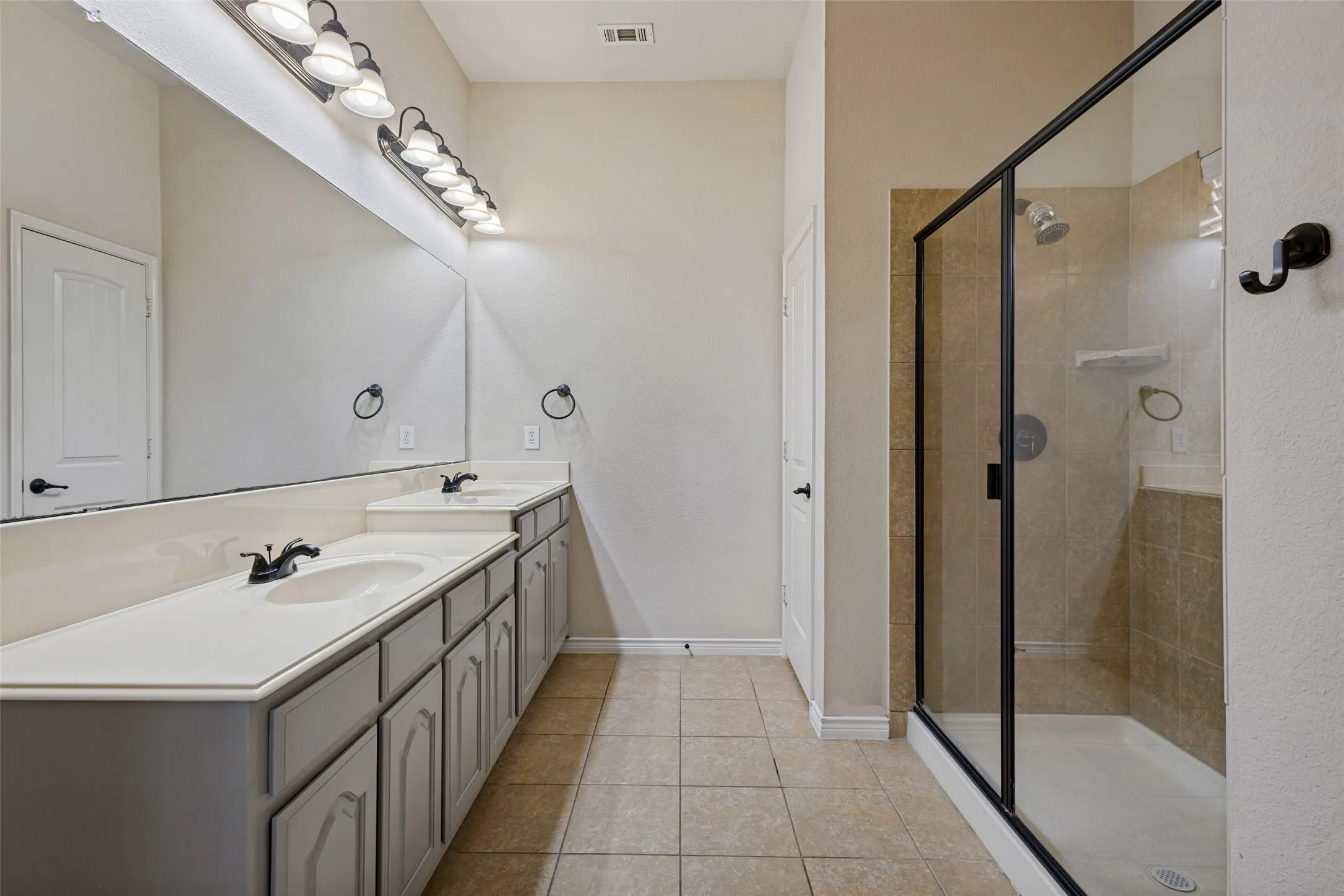 Double vanity with integrated sinks, expansive wall-mounted mirror, overhead vanity lighting, framed glass shower enclosure, and tiled shower surround
