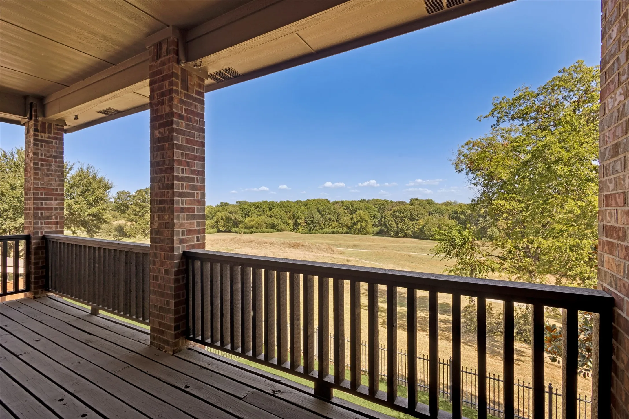 Covered outdoor balcony featuring wood decking, brick columns, and a dark wood railing