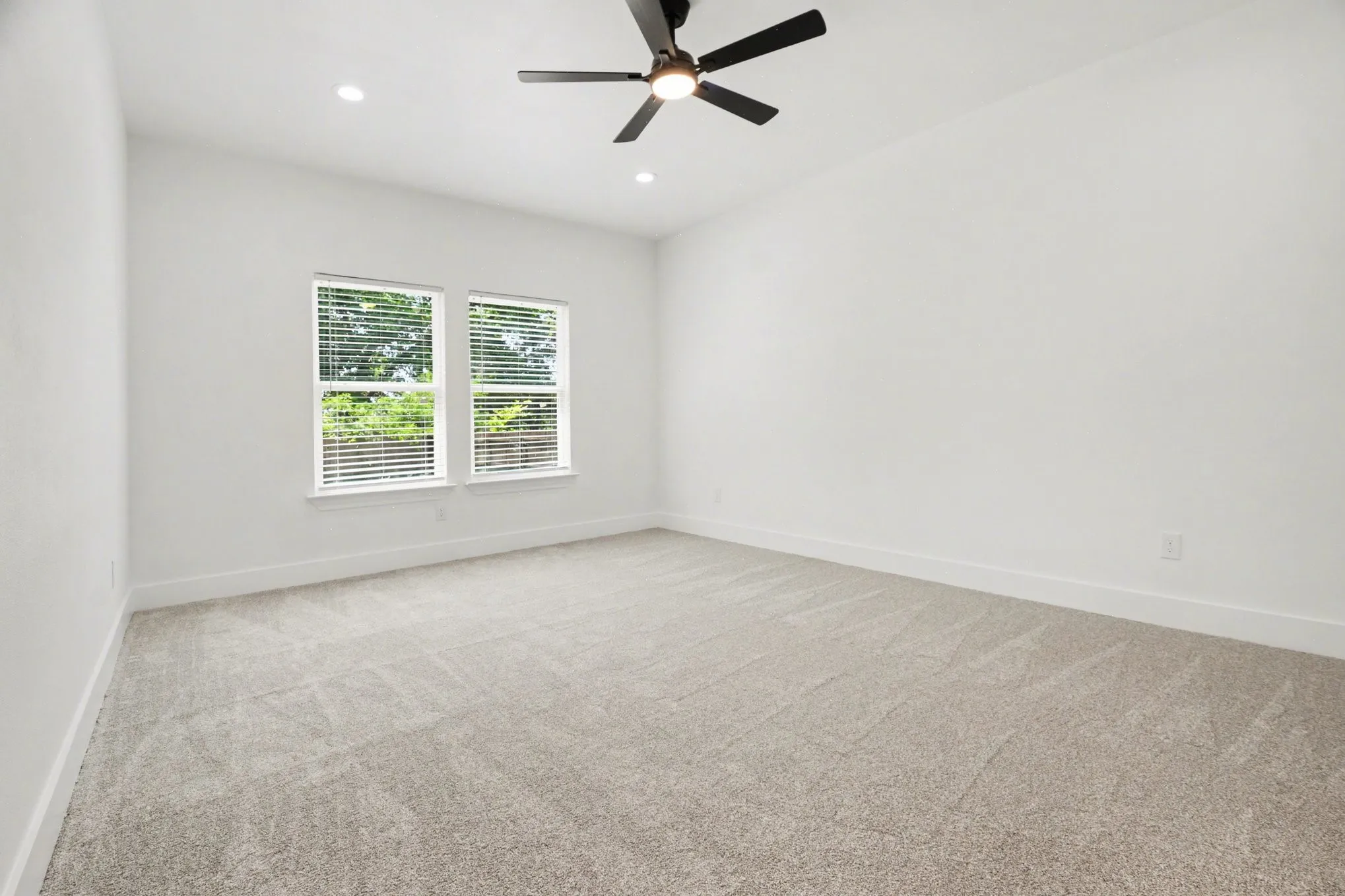 Spacious room featuring neutral carpeting, white walls, two windows with blinds, a ceiling fan, and recessed lighting